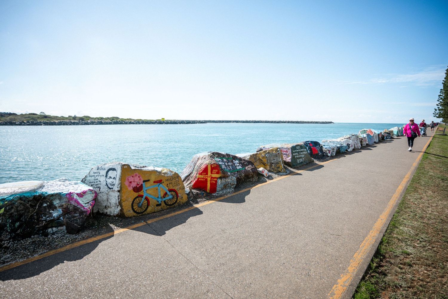 Colorful painted rocks line a coastal pathway beside turquoise water under a clear blue sky.