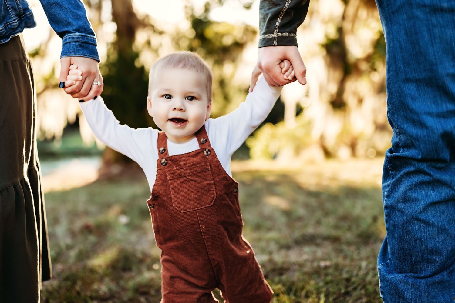 Baby in brown overalls walking while holding parents' hands in sunlit outdoor setting.