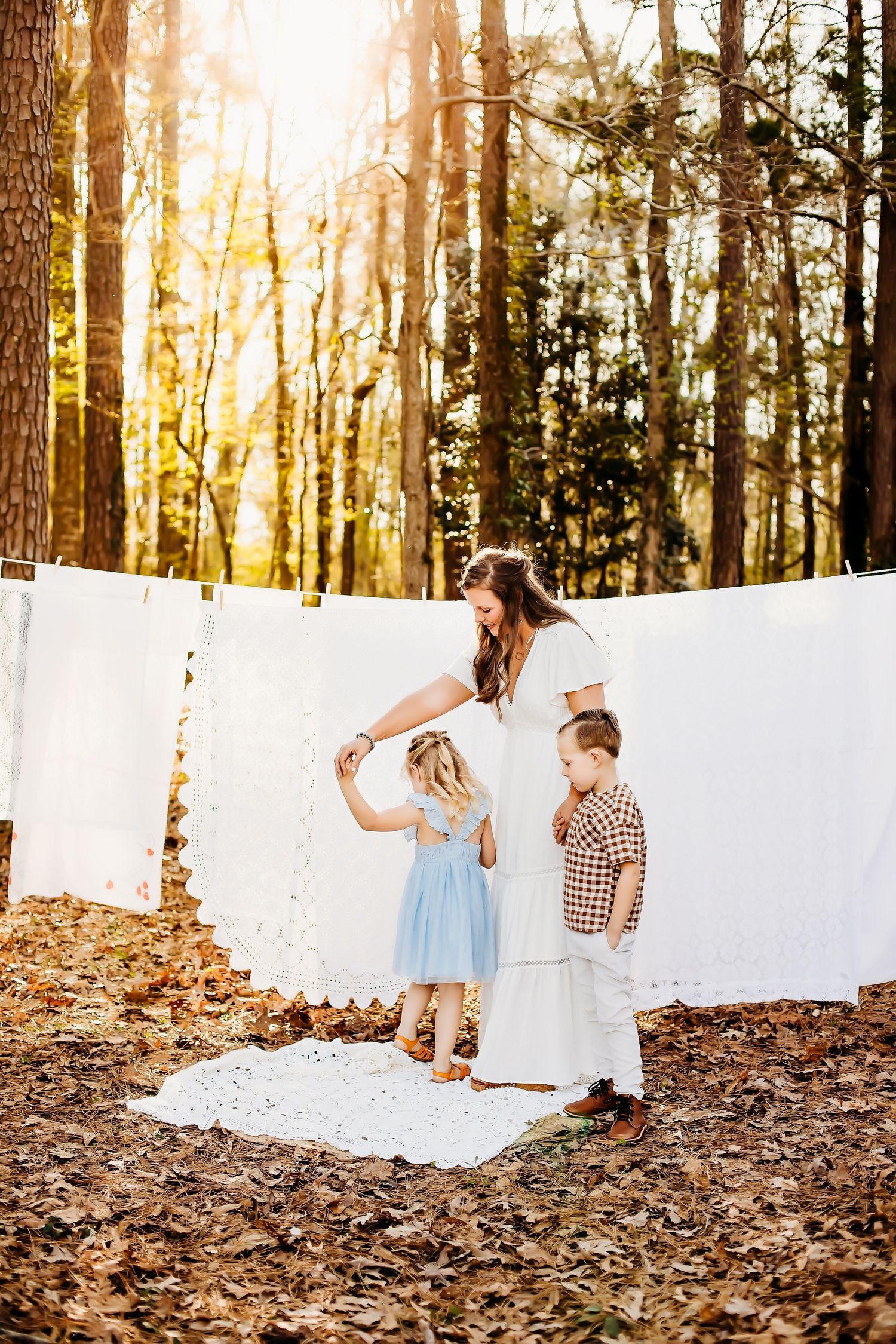 Three figures in casual wear play together against white hanging sheets in a sunlit forest.