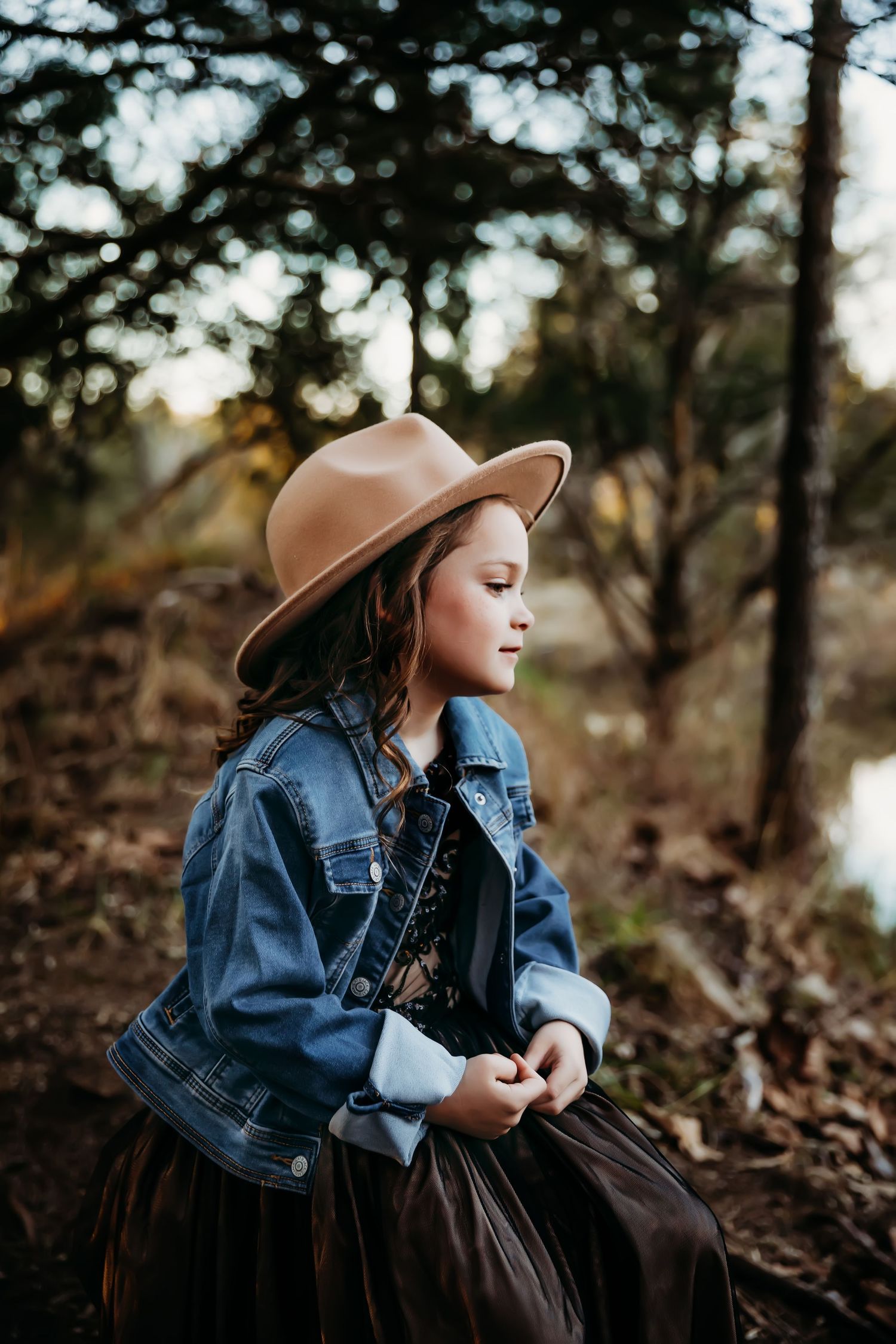 A figure in a denim jacket and brown hat sits thoughtfully by a wooded riverbank.