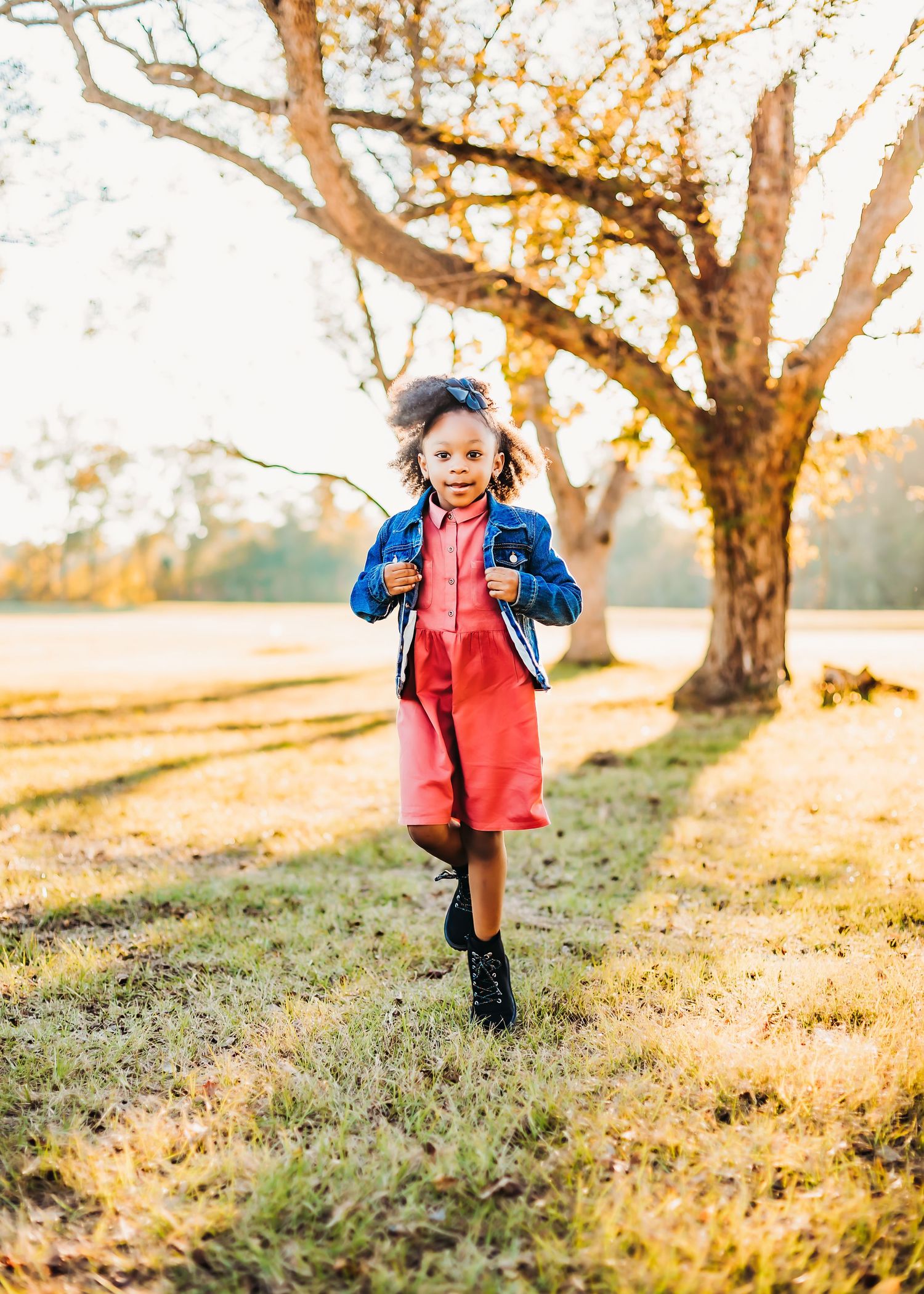A child wearing a denim jacket and red overalls runs joyfully across an autumn field at sunset.