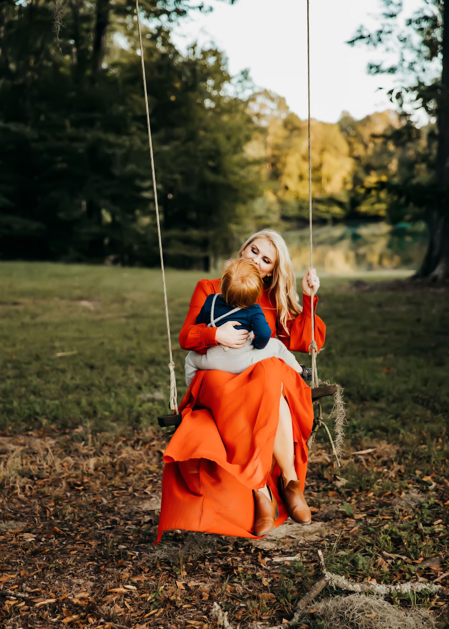 A figure in a bright orange blanket relaxes on a wooden rope swing in an autumn forest setting.