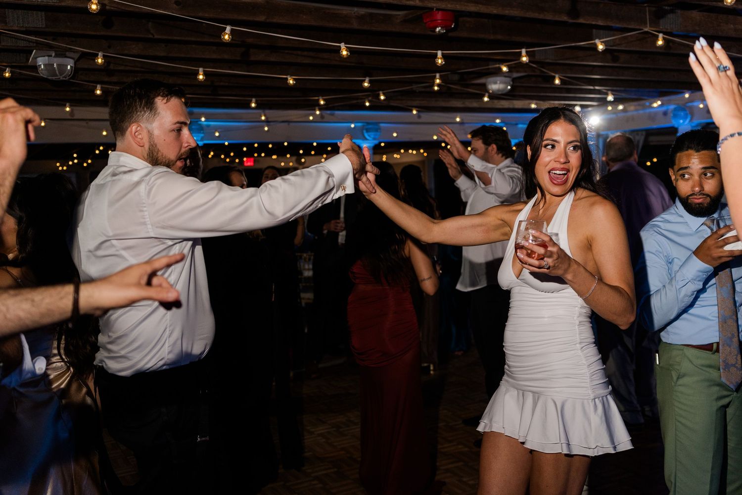 Guests dancing energetically at an indoor party with string lights, woman in white dress smiling while dancing.