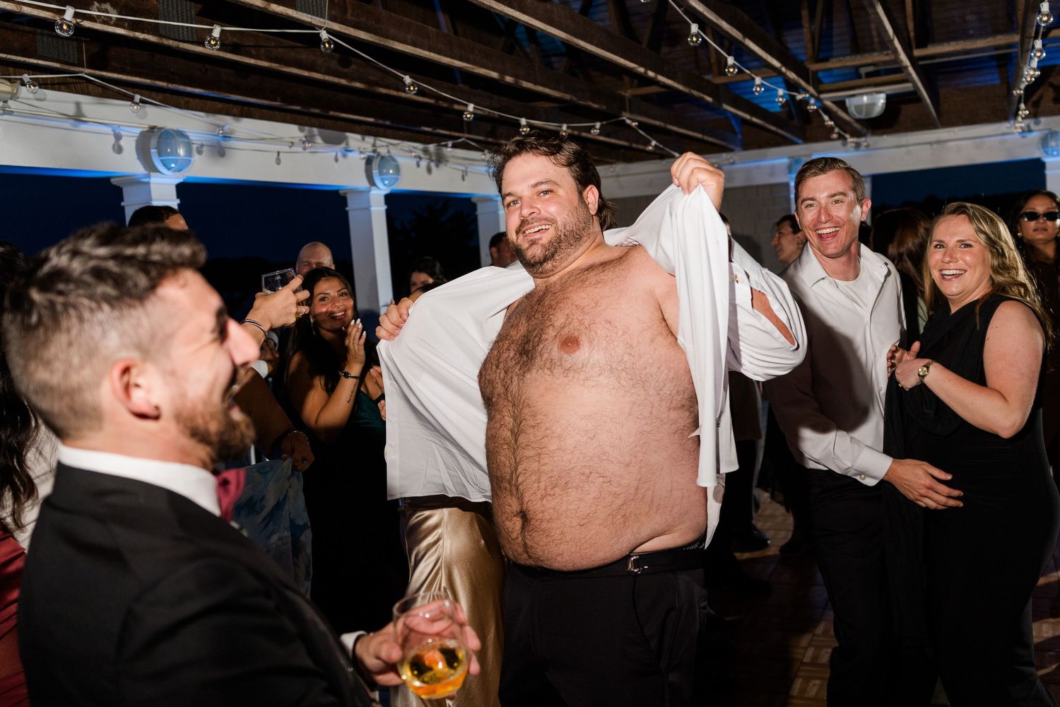 Man lifting shirt at lively party, surrounded by laughing guests in formal attire at an indoor celebration.