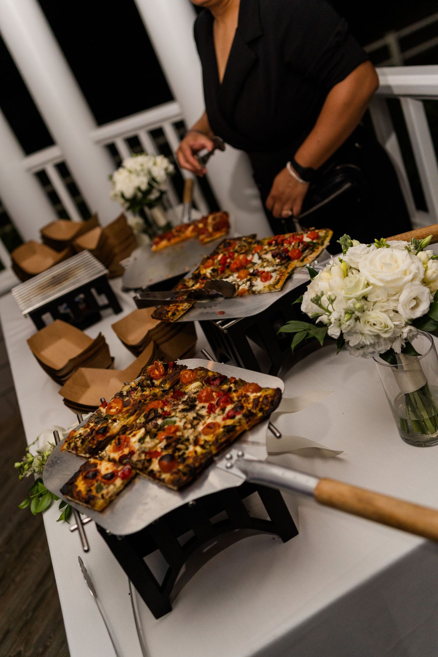 Catered event table with rectangular pizzas topped with vegetables, served with spatulas and white floral arrangements.