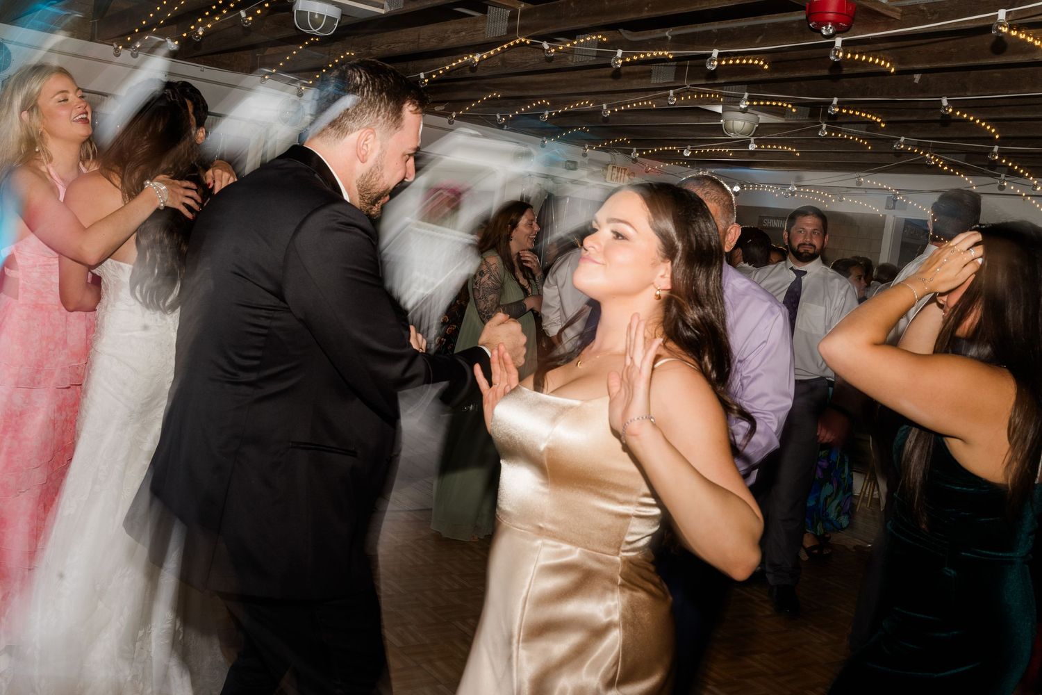 Guests dancing joyfully at a wedding reception, with the bride in a gold satin dress and groom in a black tuxedo.