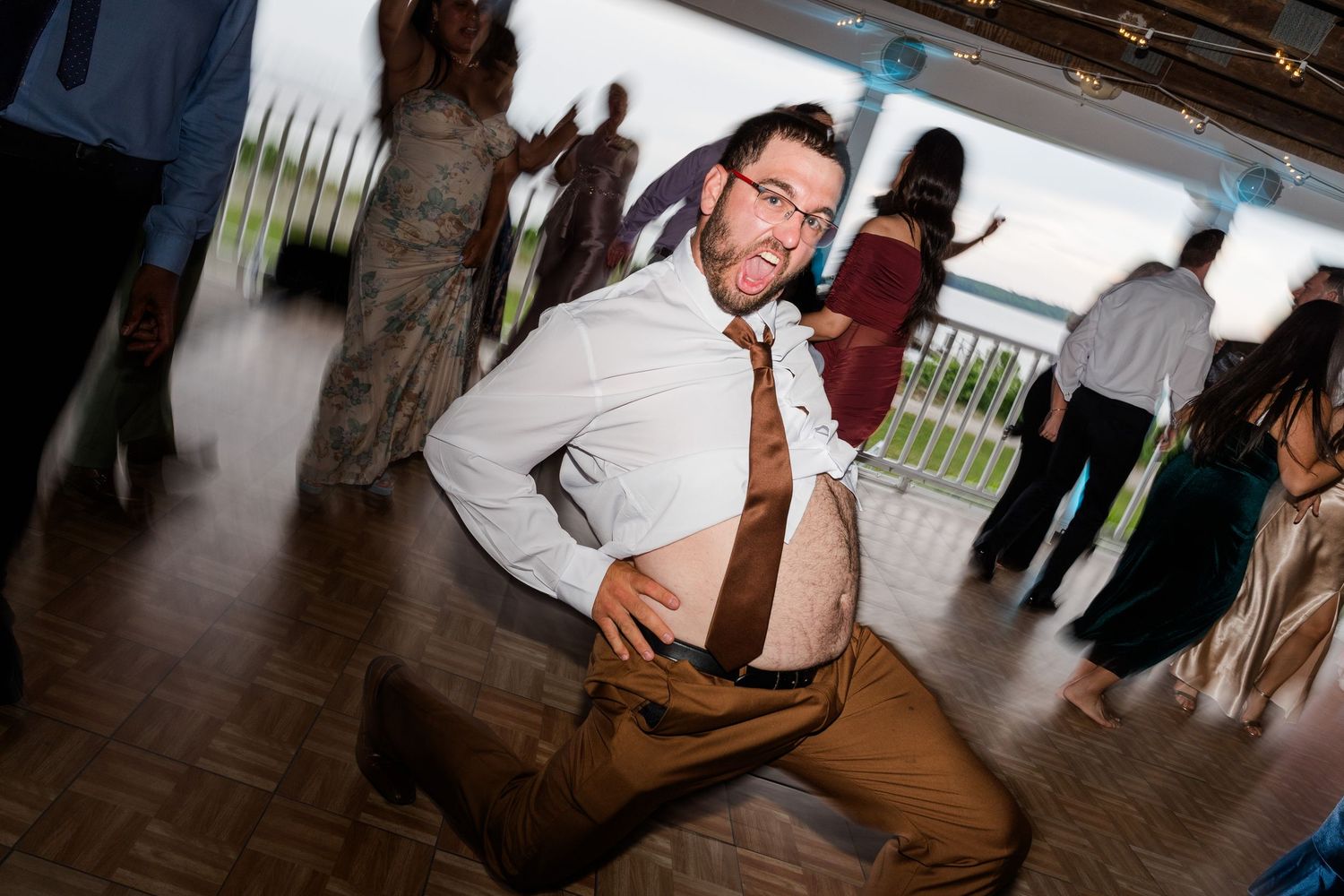 Man dancing energetically at a wedding reception, kneeling on the dance floor in a white shirt and loosened tie.