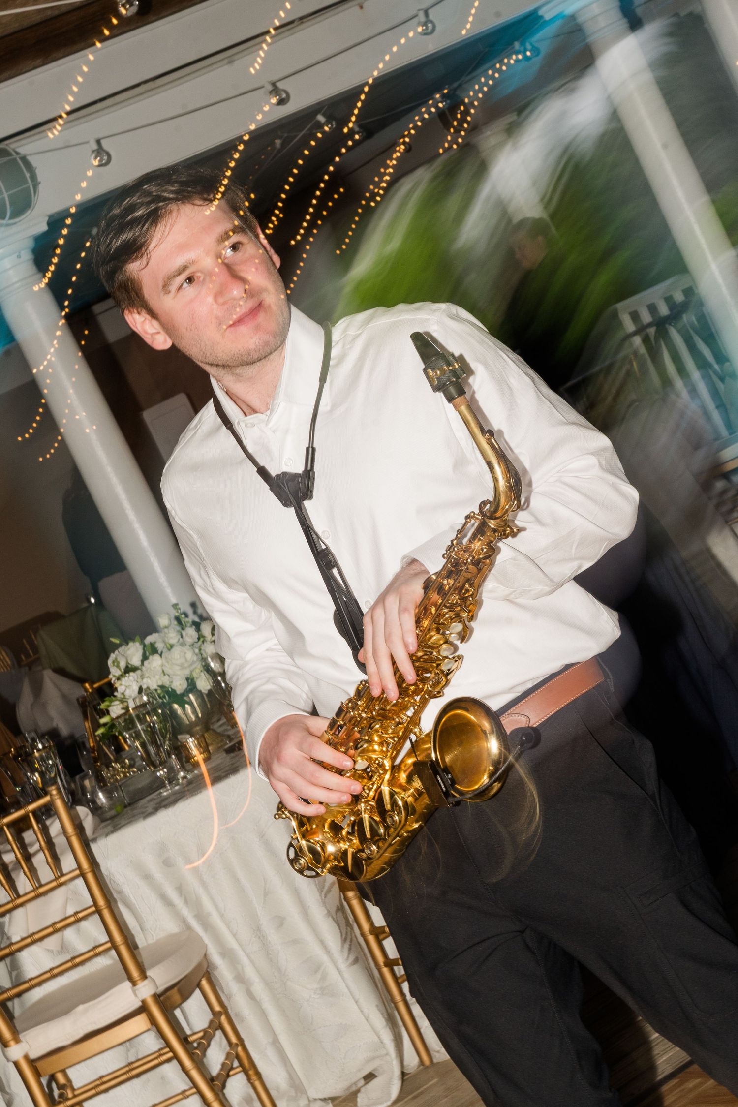 A saxophonist in a white shirt plays alto saxophone at an elegant wedding reception with string lights.
