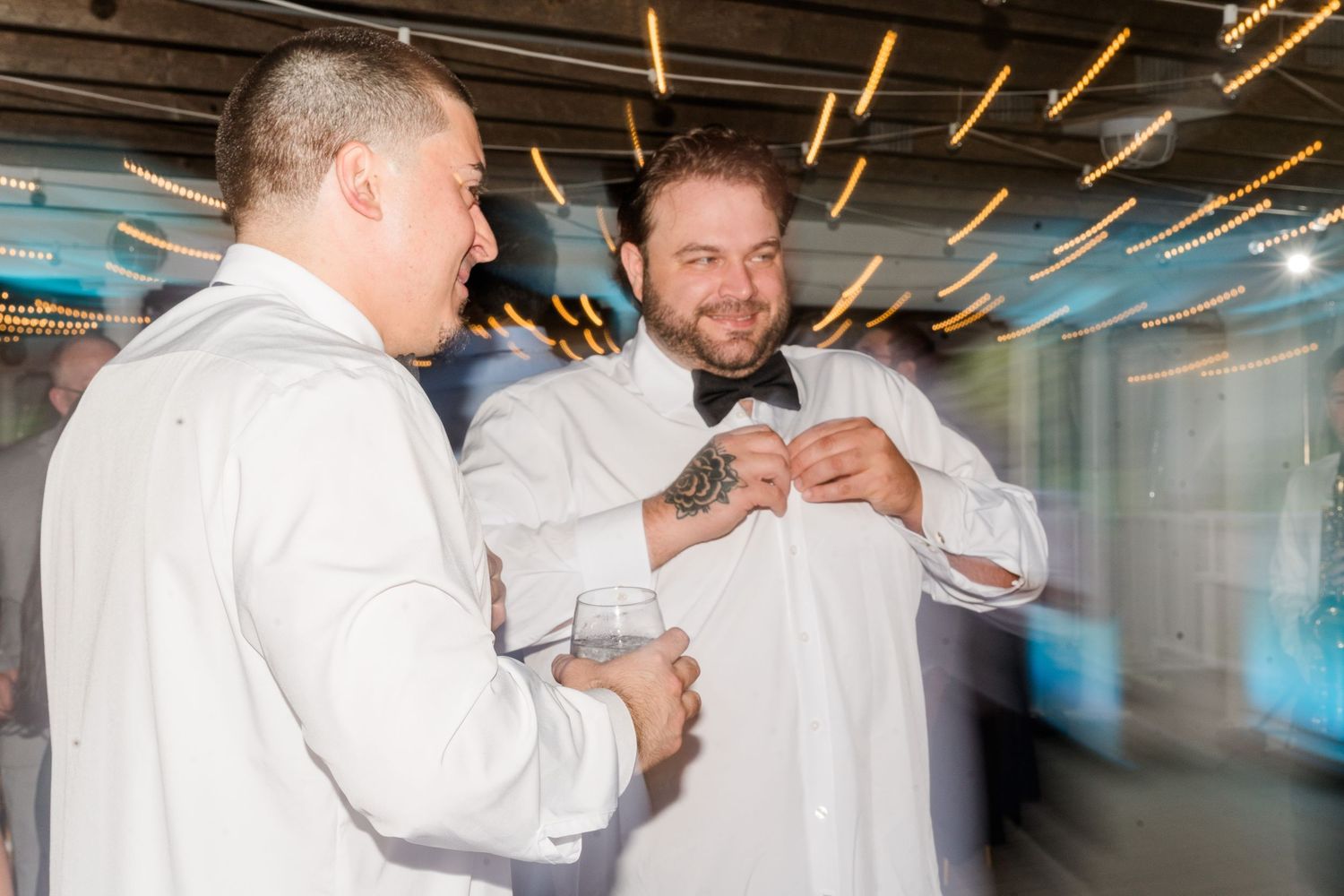 Two men in white dress shirts laughing and sharing a drink at an elegant wedding reception.