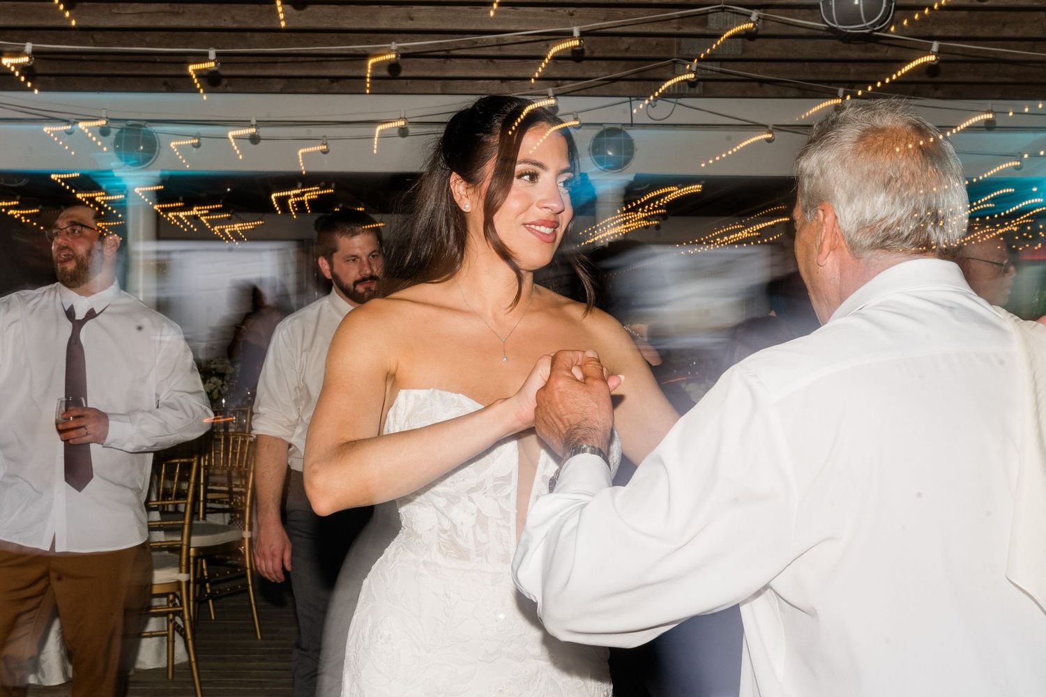 Bride in white gown smiling while dancing with guest at elegant wedding reception with string lights.