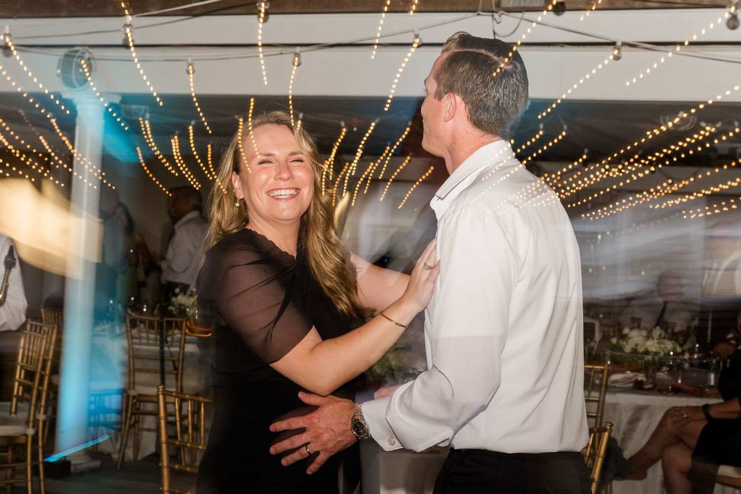 Couple dancing and smiling together at a wedding reception under glowing string lights.