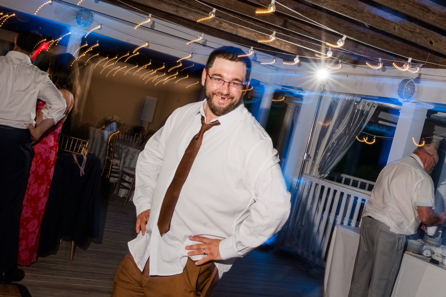 Man in white shirt and brown tie smiling on wedding reception dance floor with string lights.
