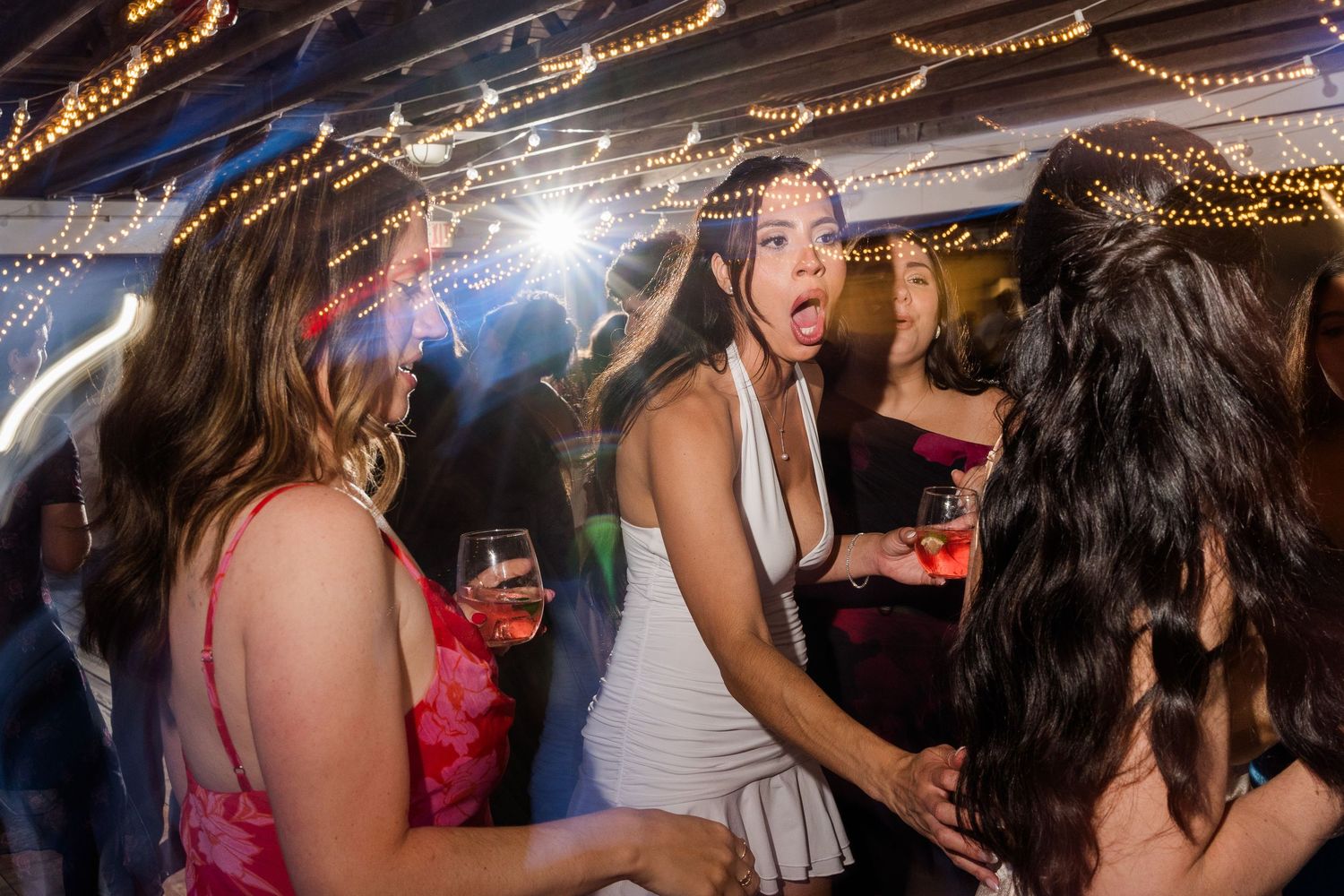Excited women dancing and celebrating at a festive wedding reception party under warm string lights.