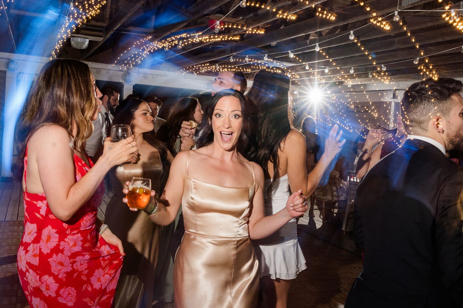 Joyful woman in gold satin dress dancing at a lively wedding reception with string lights and guests.