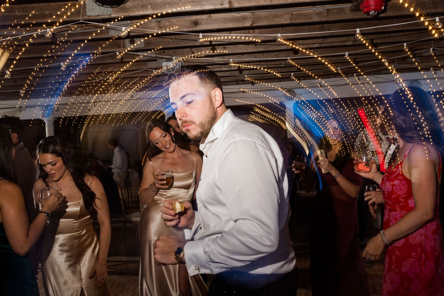 Couple slow dancing at a wedding reception under string lights with guests in background.