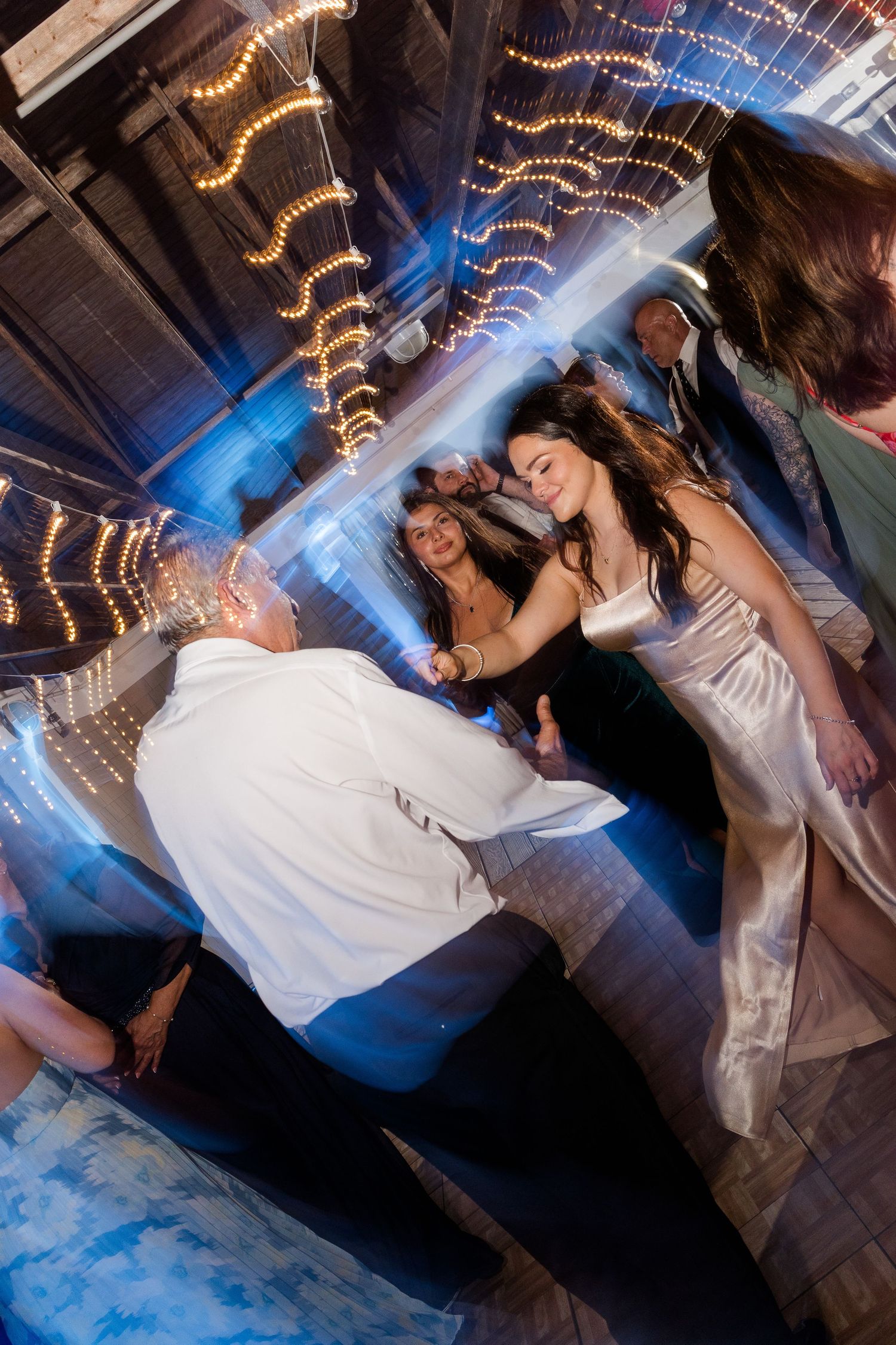 Bride and groom dancing energetically at wedding reception with string lights and blue lighting.
