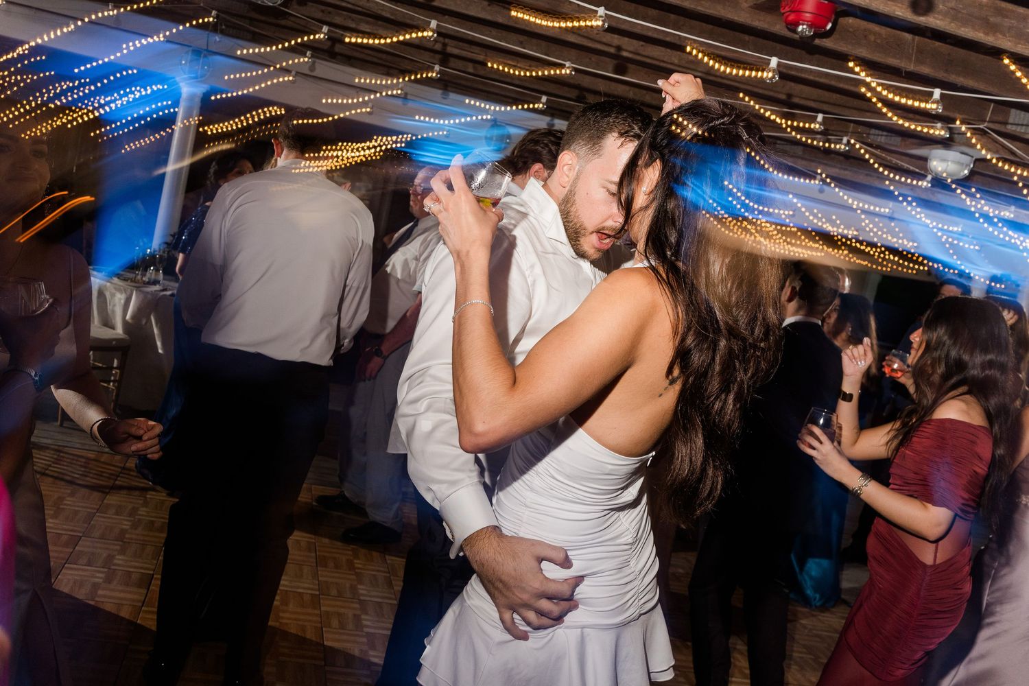 Bride and groom share romantic first dance at wedding reception with guests dancing in background.