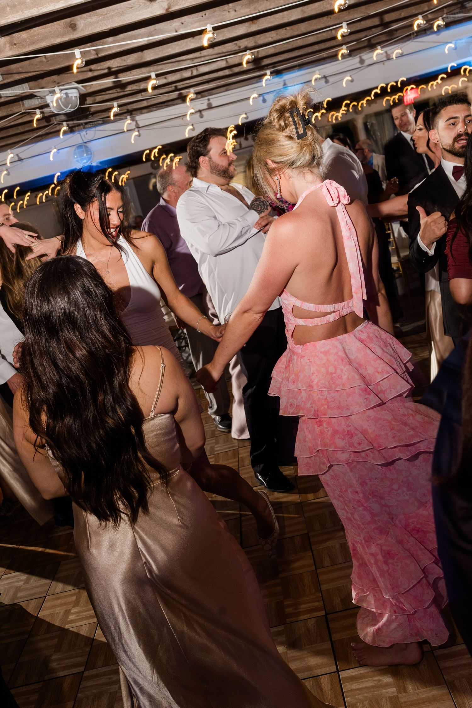 Guests dancing at a formal event, women in elegant gowns including a pink ruffled backless dress on a wooden dance floor.