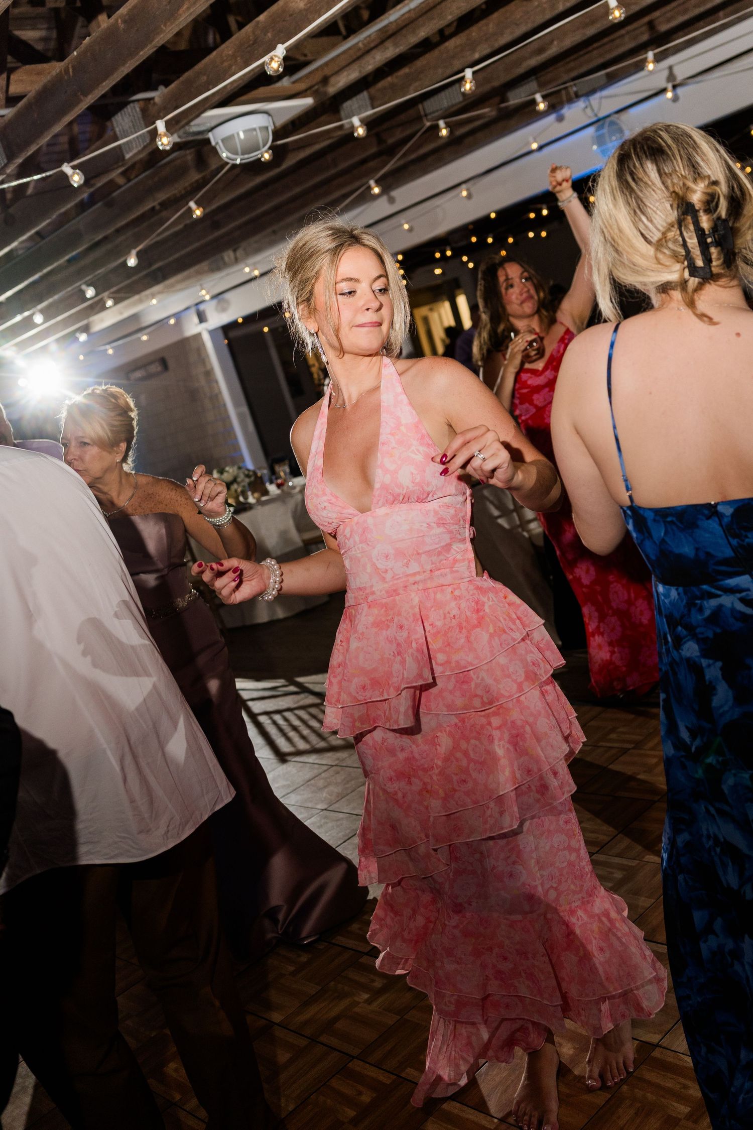 Woman in pink ruffled halter dress dancing at an elegant indoor wedding reception with string lights.