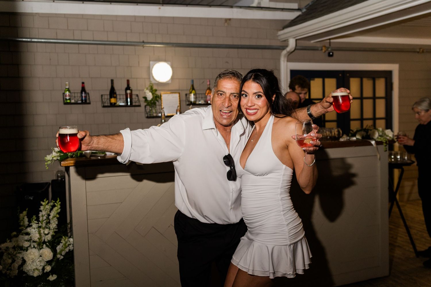 Smiling couple posing with drinks at a wedding reception bar, man in white shirt and woman in white dress.