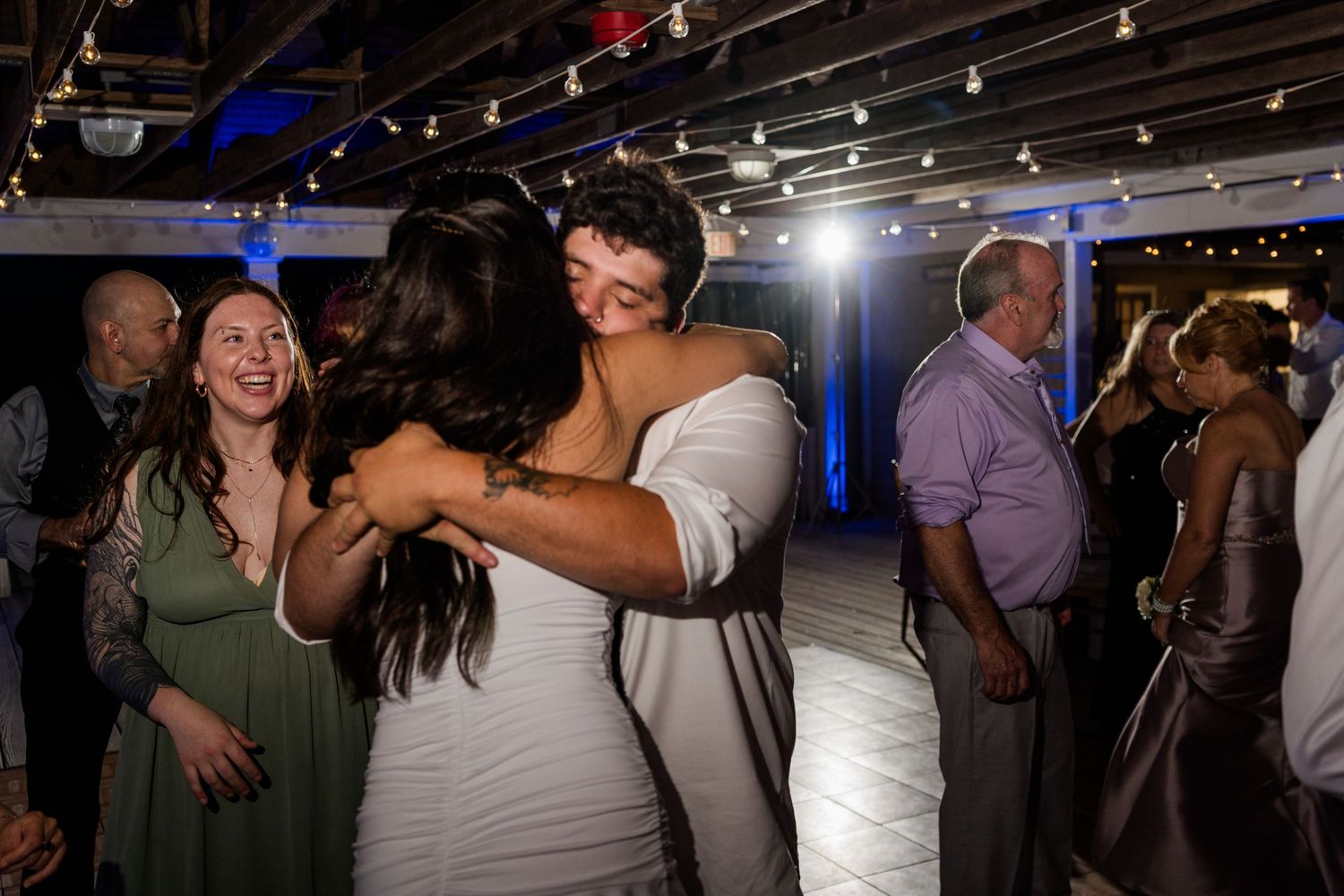 Two brides embrace joyfully on a decorated wedding reception dance floor surrounded by smiling guests.