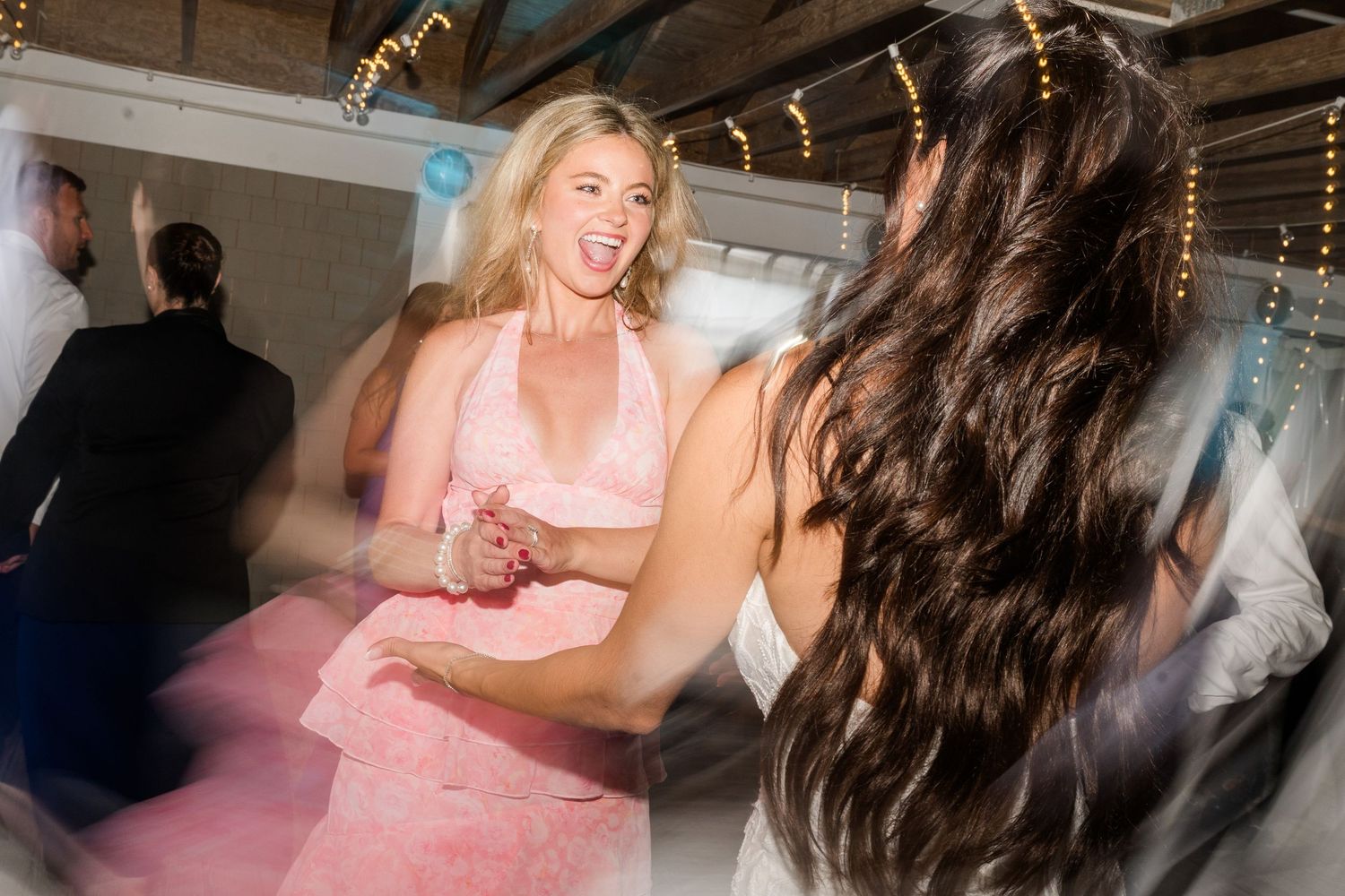 Two women in formal dresses dancing and laughing joyfully at an elegant indoor event or wedding reception.