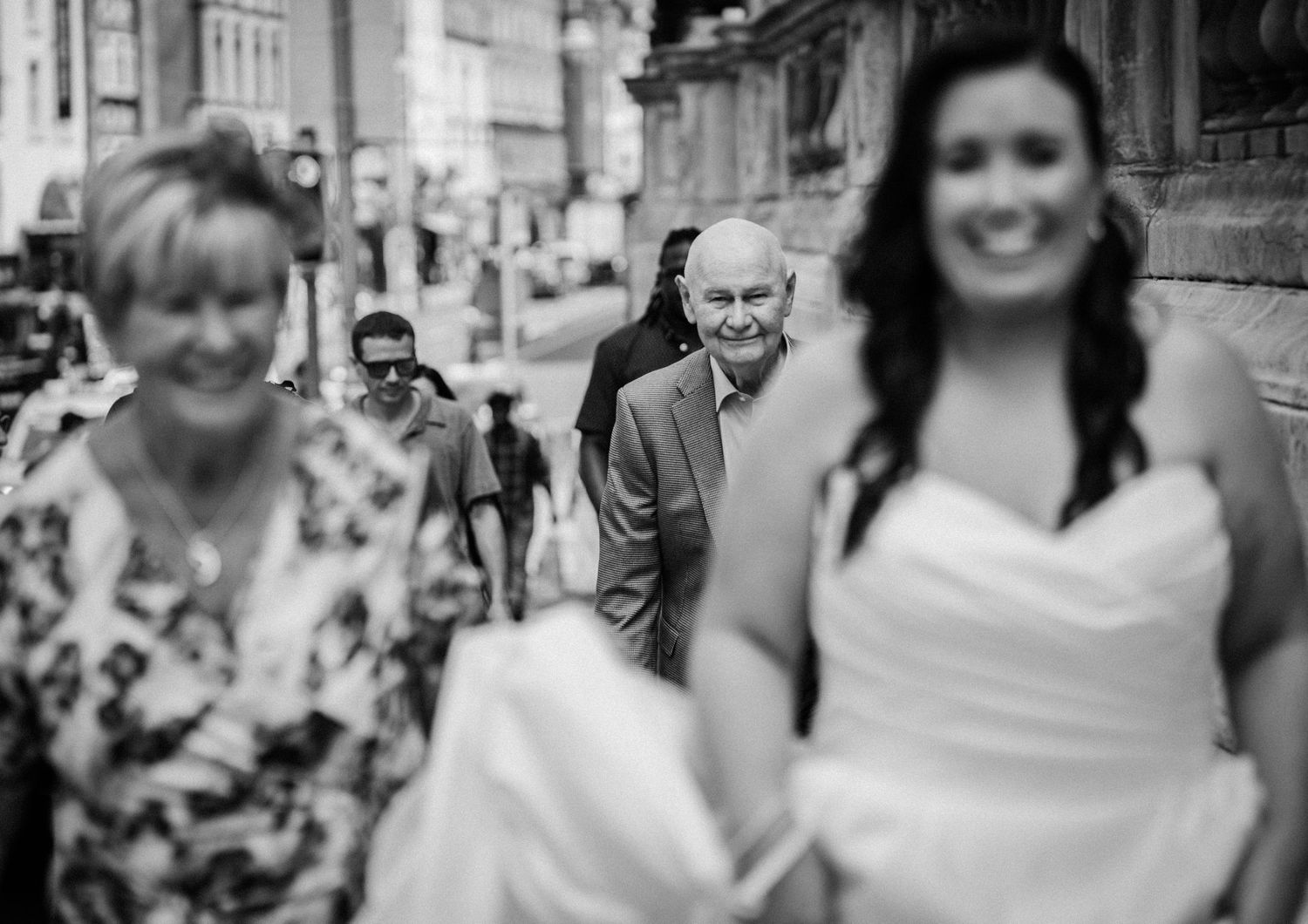 Black and white wedding photo of a smiling bride in white dress walking with family on a city street.