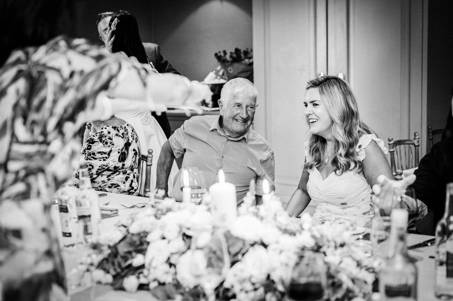 Black and white wedding reception photo of guests laughing at a decorated table with candles and floral centerpieces.