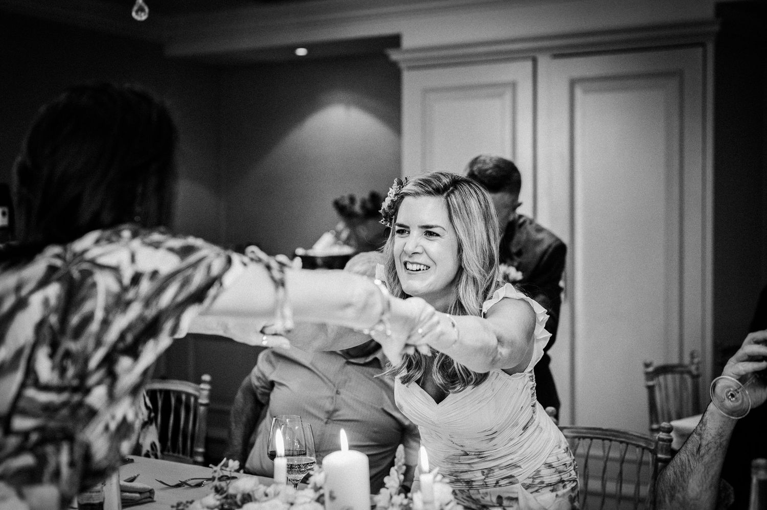 Black and white photo of a woman laughing and reaching across a wedding reception table with candles and floral centerpieces.