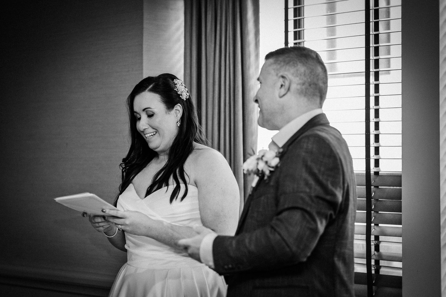 Black and white photo of a bride reading wedding vows while smiling, standing beside her groom near a window.
