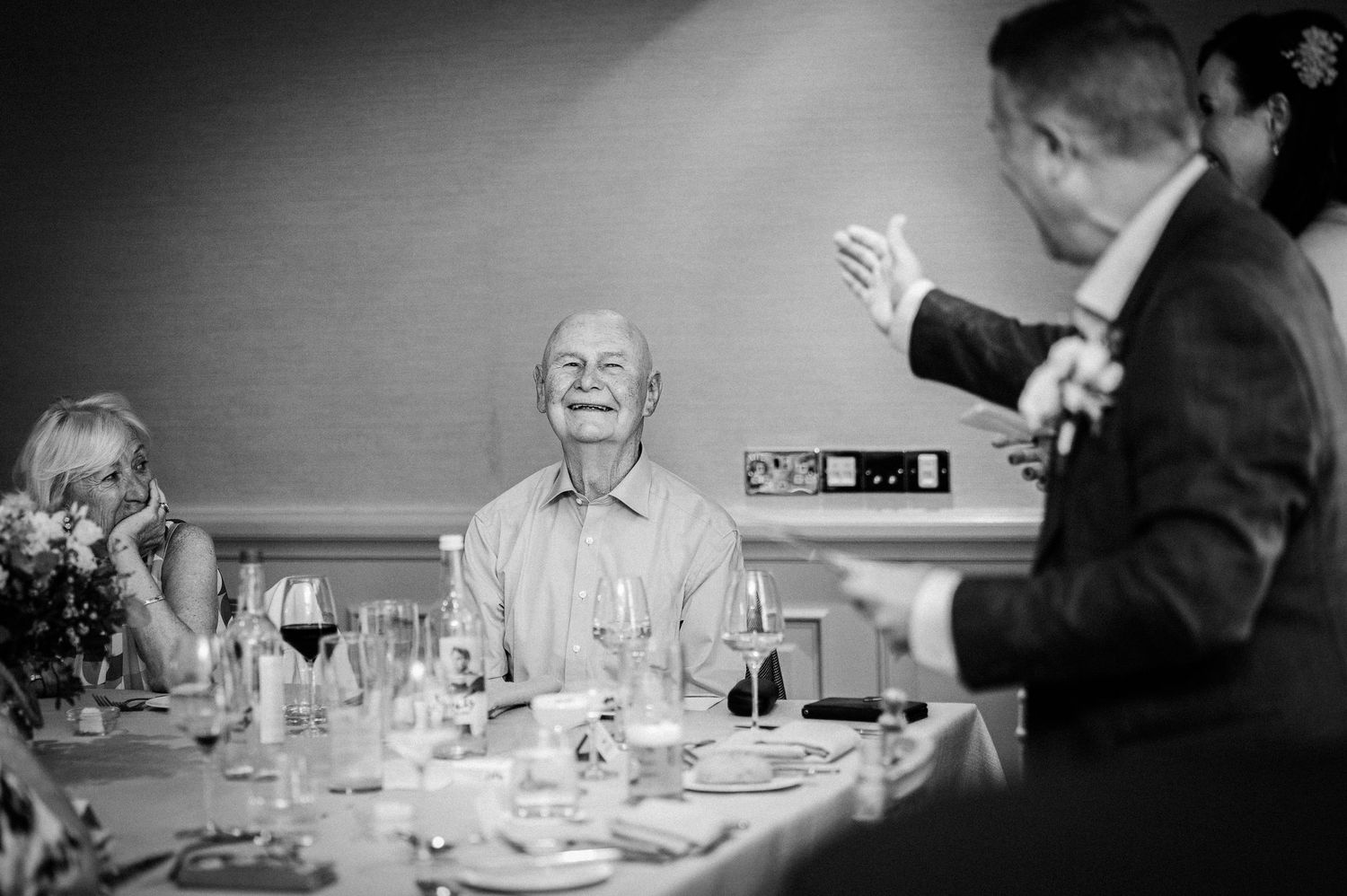 Elderly man laughing joyfully at a wedding reception dinner table in black and white photography.