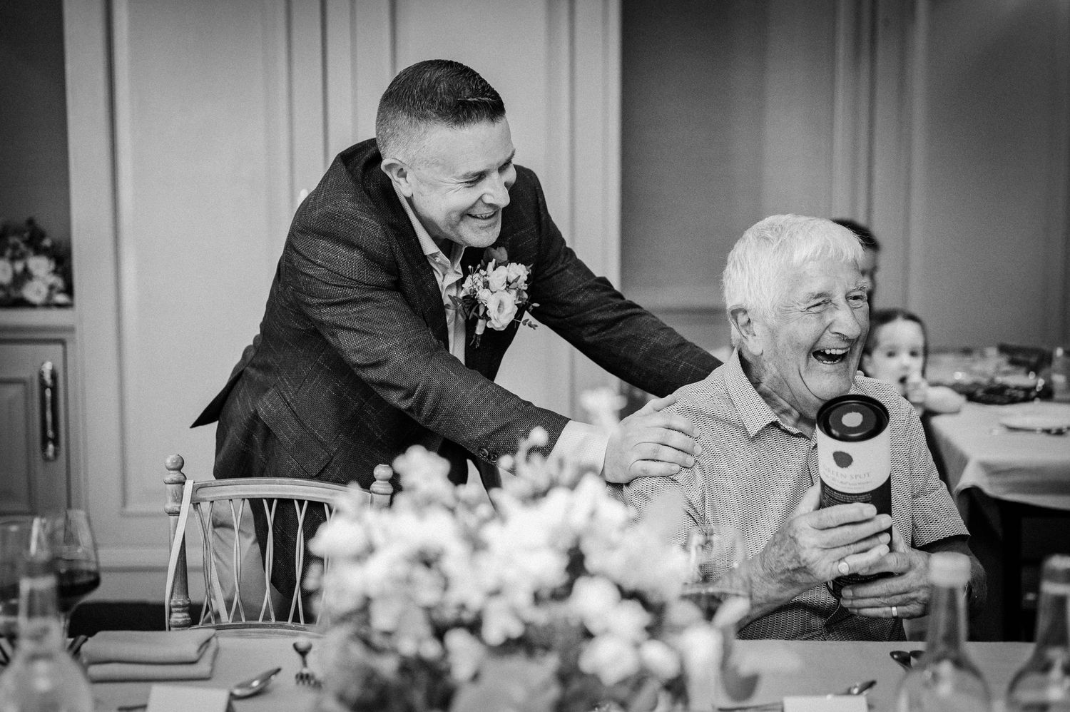 Two men laughing joyfully at a wedding reception, one holding a gift, captured in elegant black and white.
