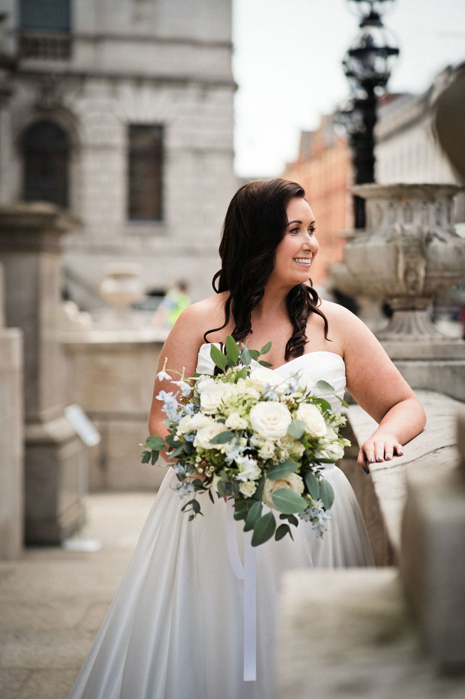 Smiling bride in strapless white gown holding lush white and green bouquet, posing on stone steps at an urban venue.