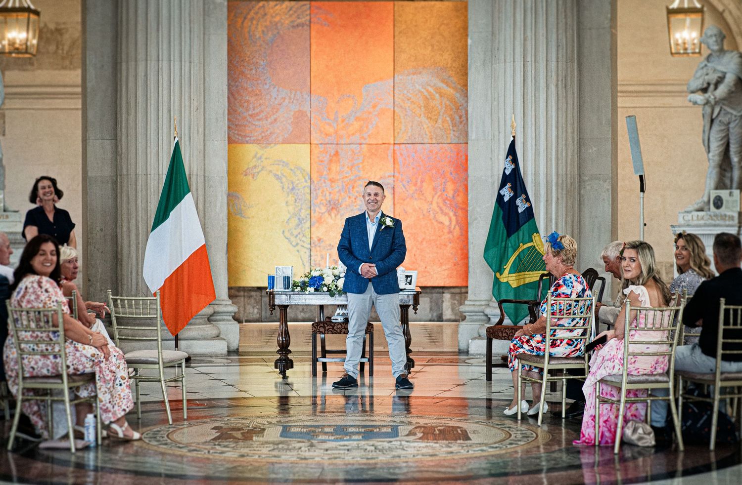 Man speaking at elegant hall with Irish flag, seated guests in colorful attire, and vibrant mural backdrop.