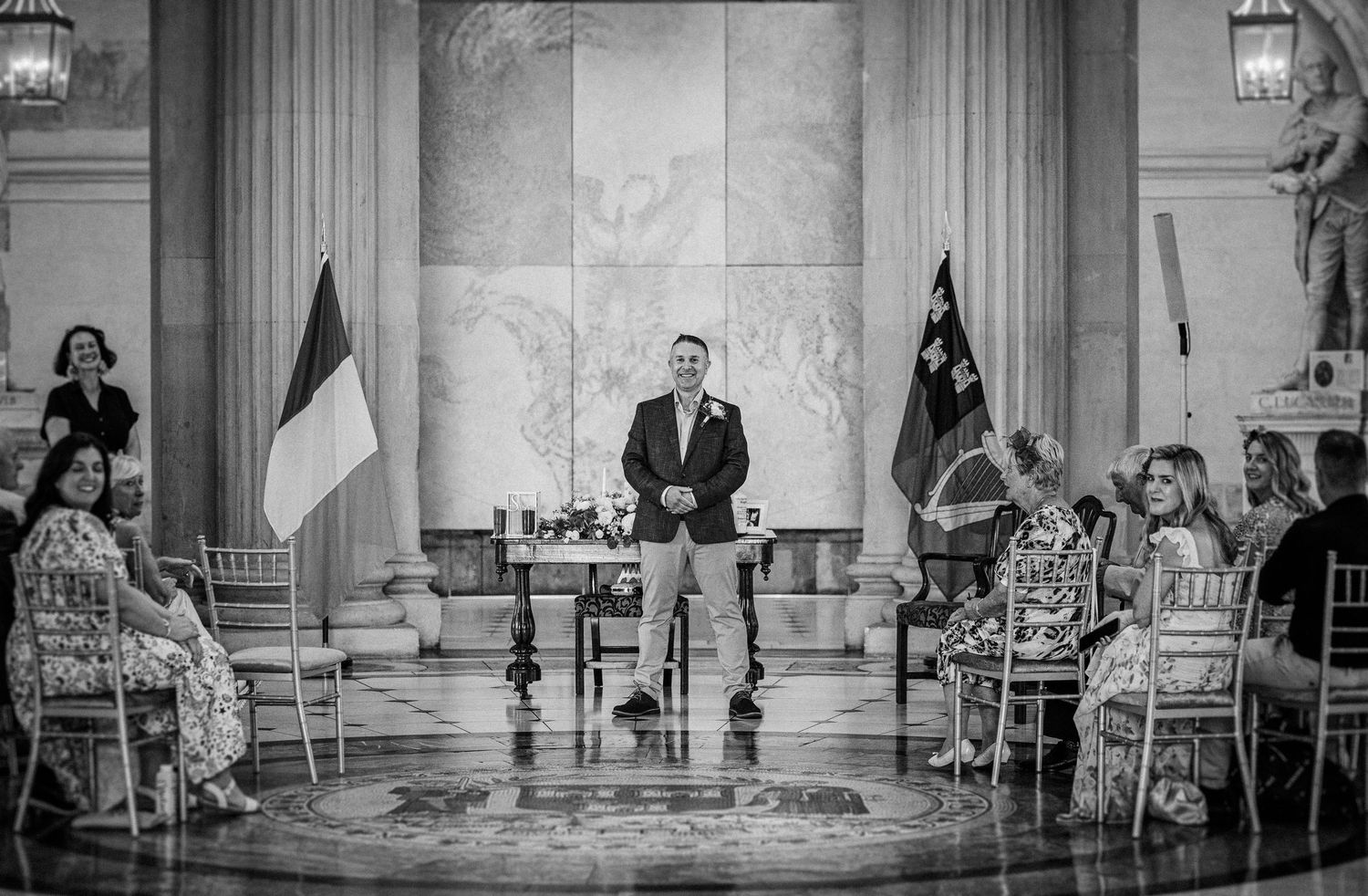 Black and white wedding ceremony photo in an elegant hall with guests seated and officiant standing between two flags.