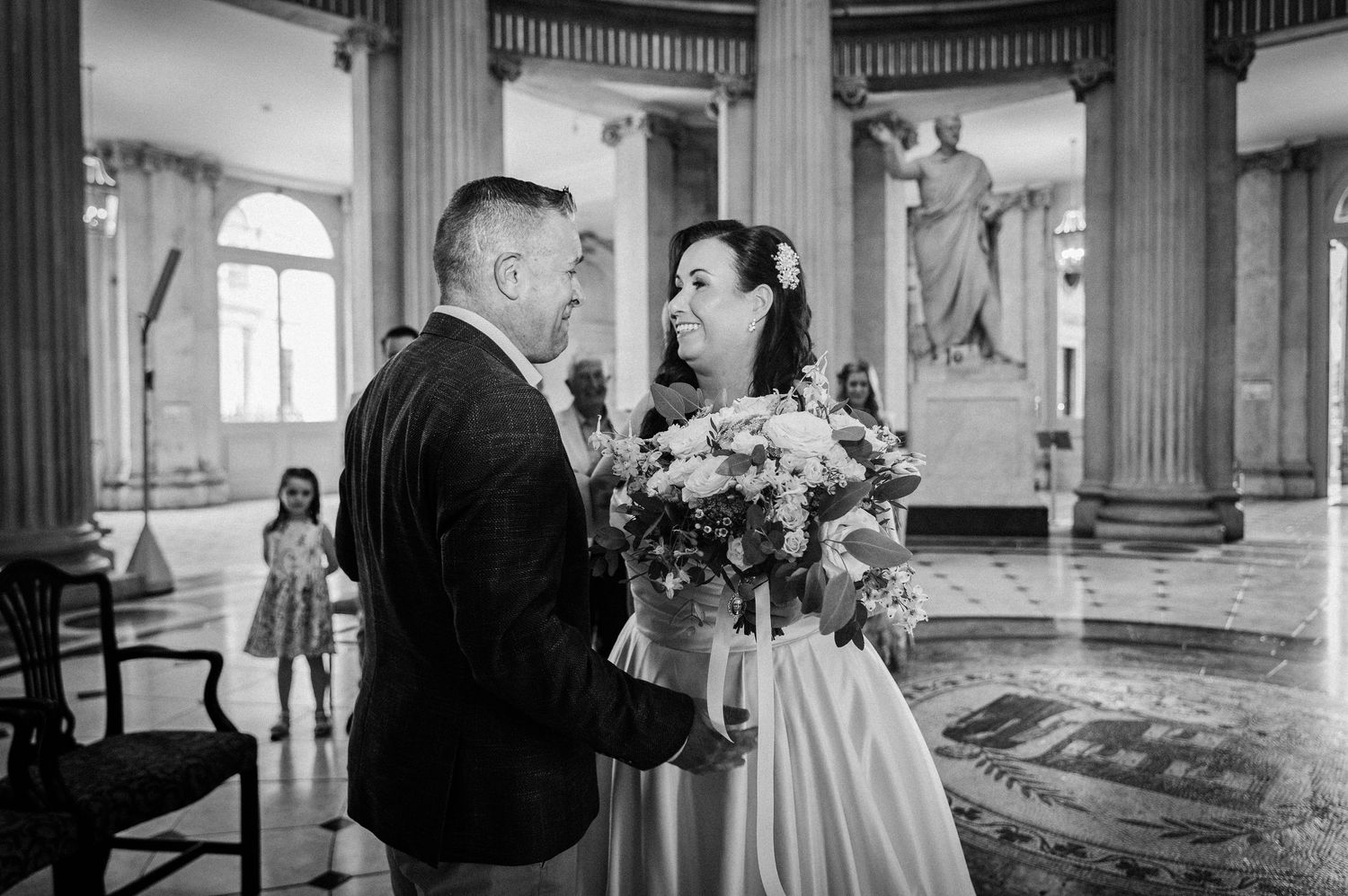 Black and white photo of bride and groom sharing a tender moment in a grand classical hall with statue.