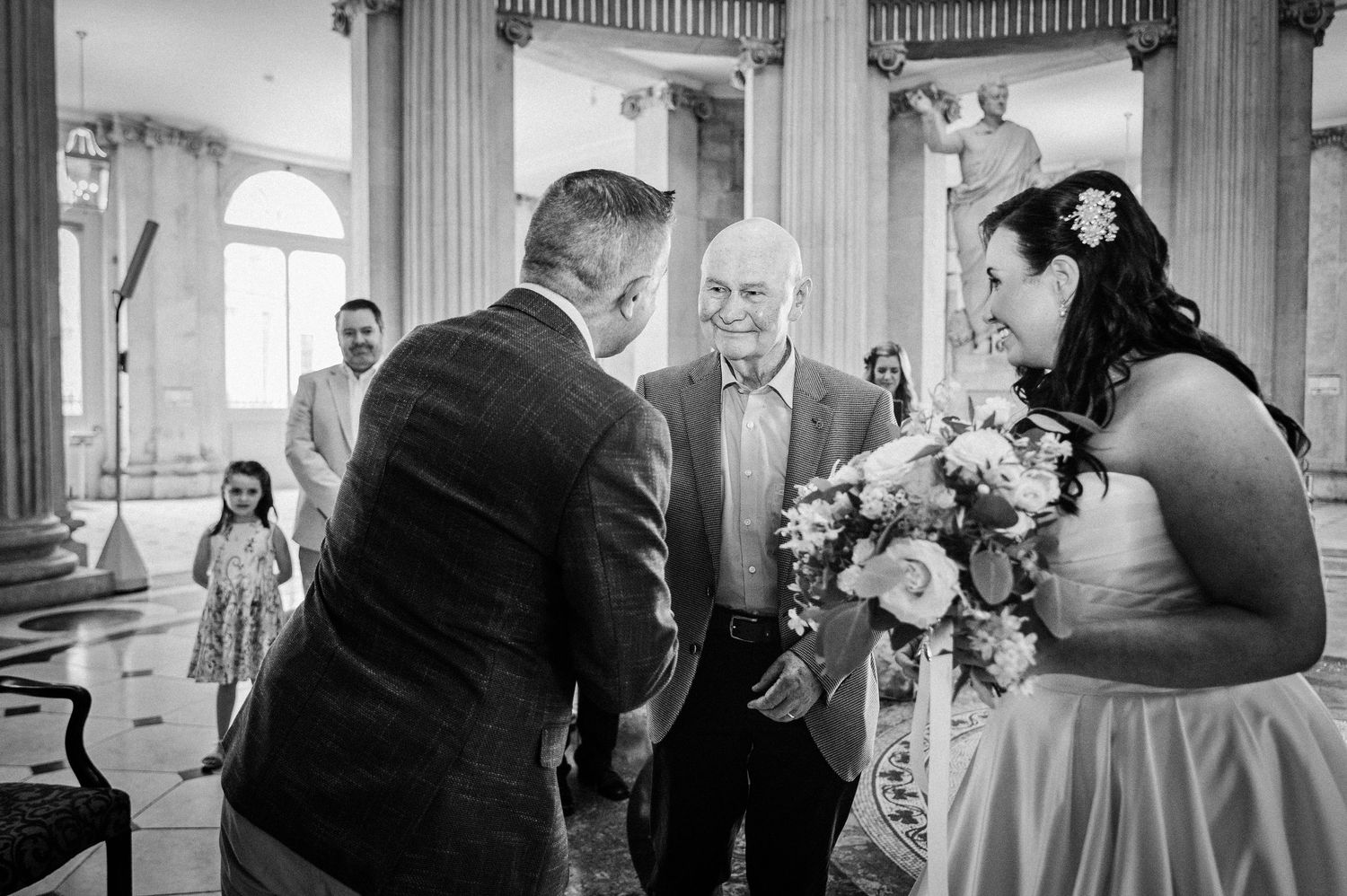Black and white wedding photo of bride holding bouquet greeting elderly guest in elegant venue.