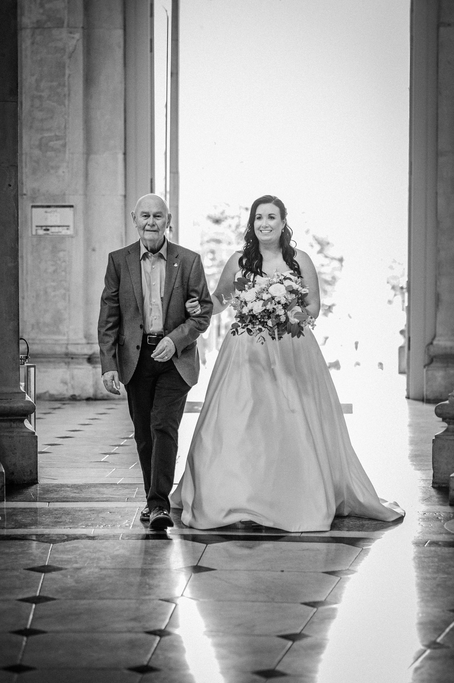 Black and white photo of a bride in a ball gown holding flowers, walking down the aisle with an older man.