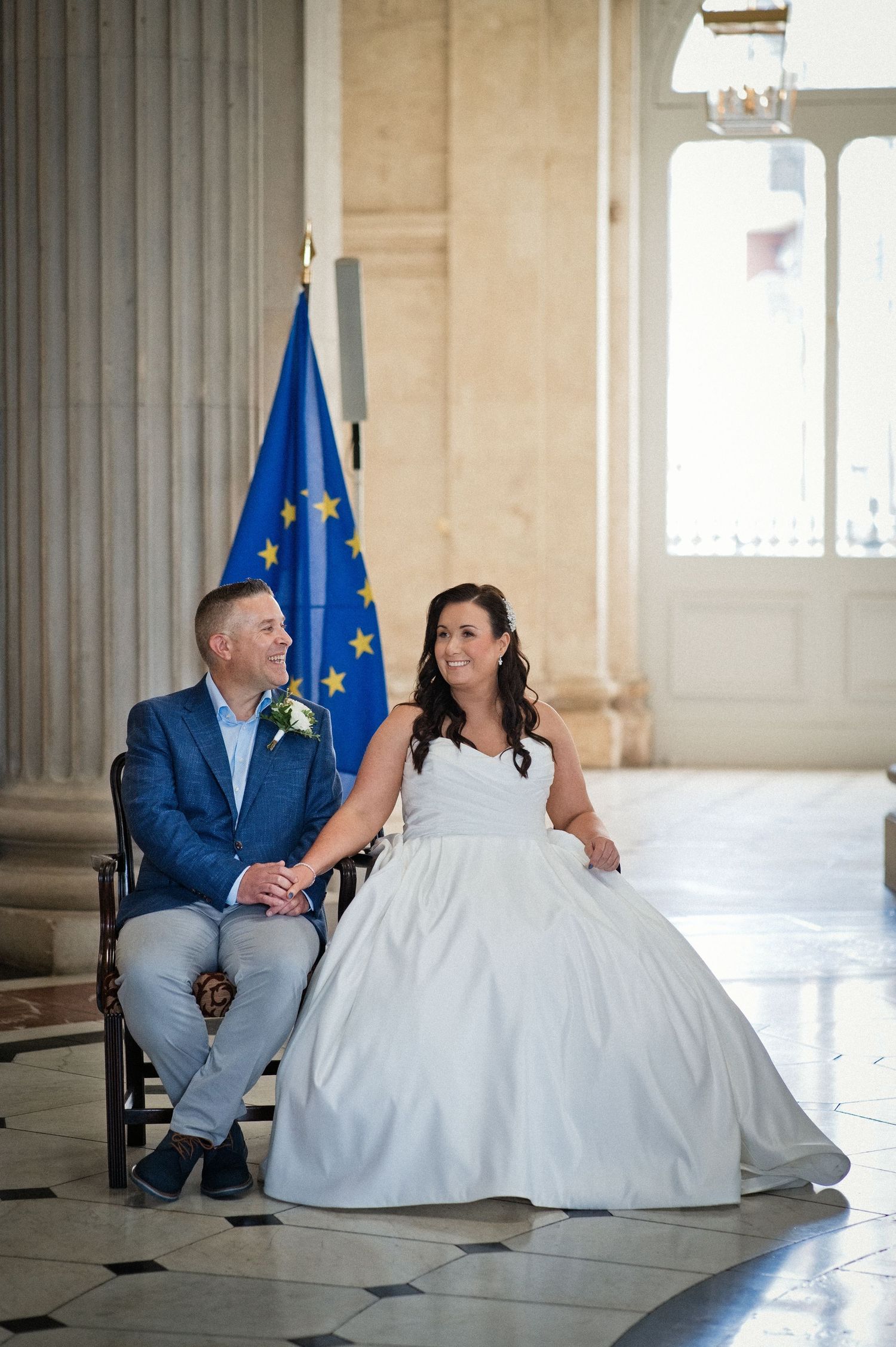 Couple in wedding attire sitting together in elegant hall with EU flag, bride in white gown, groom in blue suit.