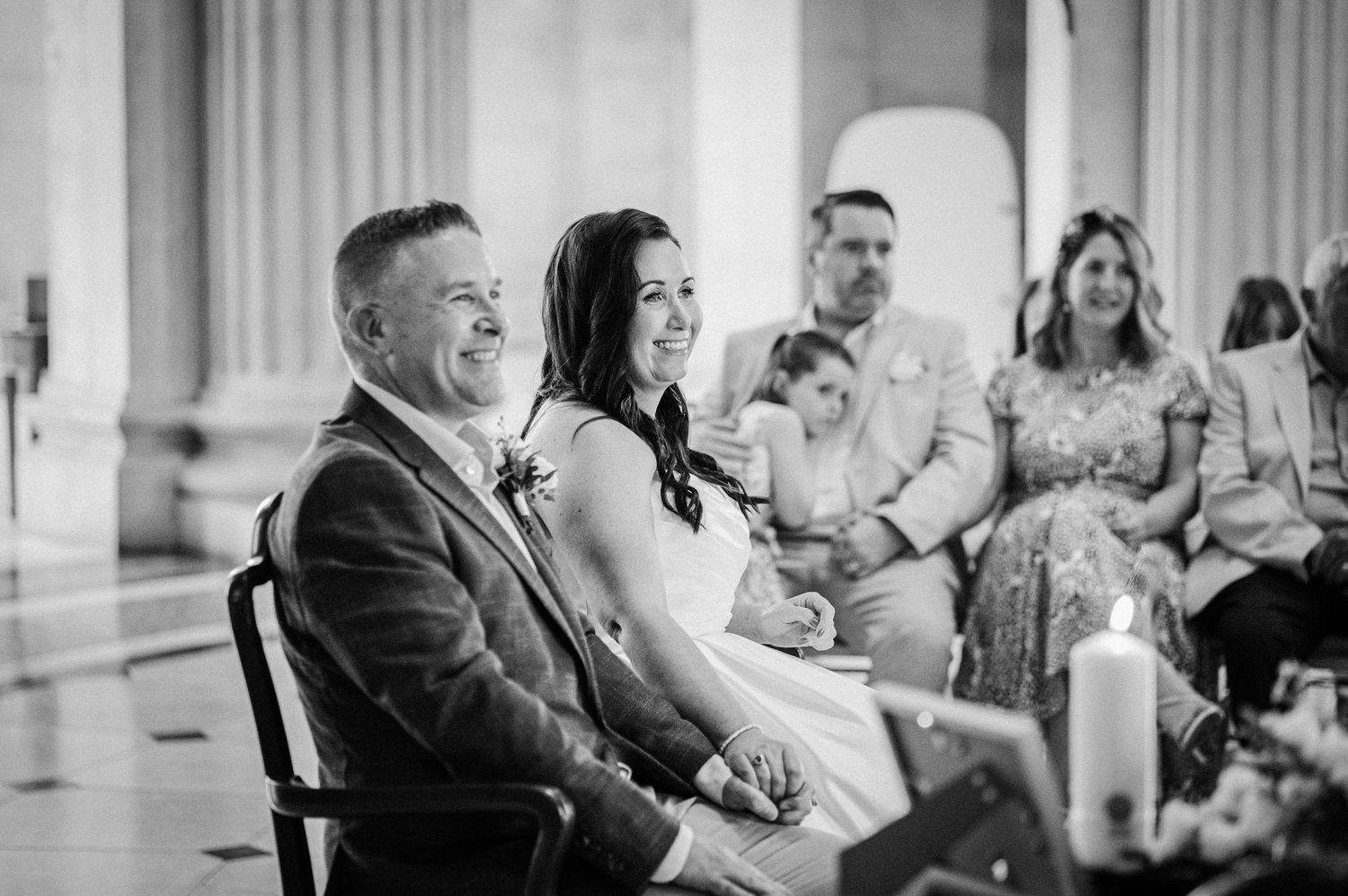 Black and white photo of a smiling bride and groom seated at their wedding ceremony with guests in the background.