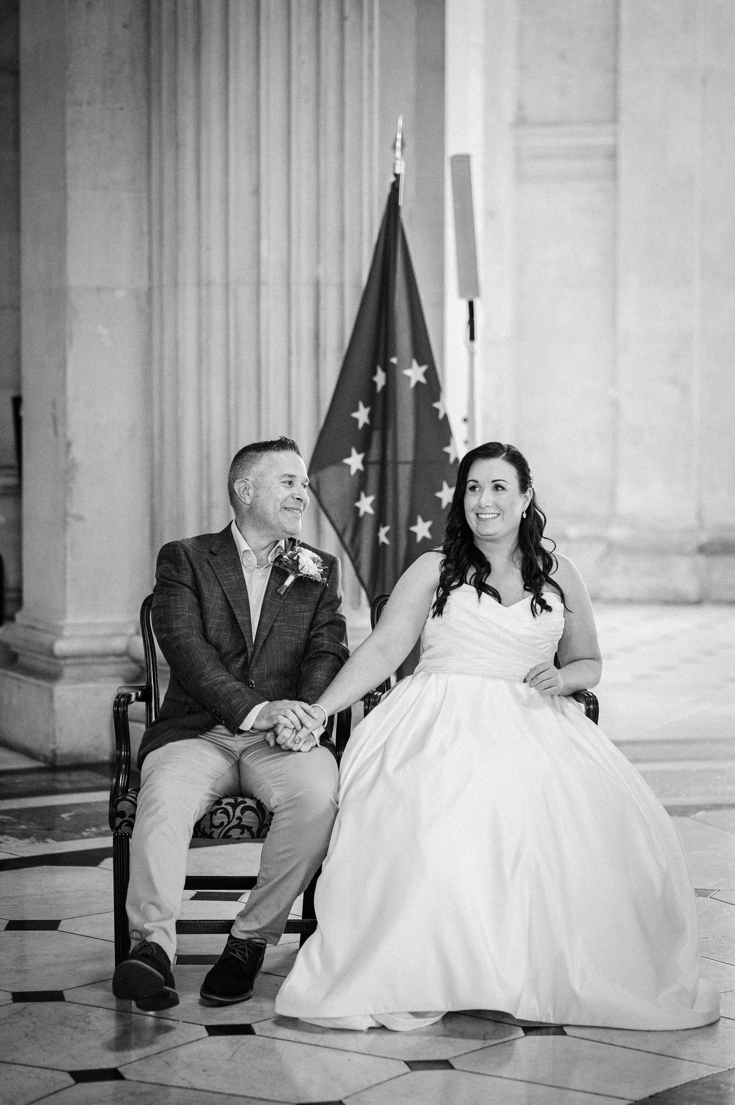 Black and white photo of bride and groom holding hands seated before EU flag at elegant wedding ceremony.
