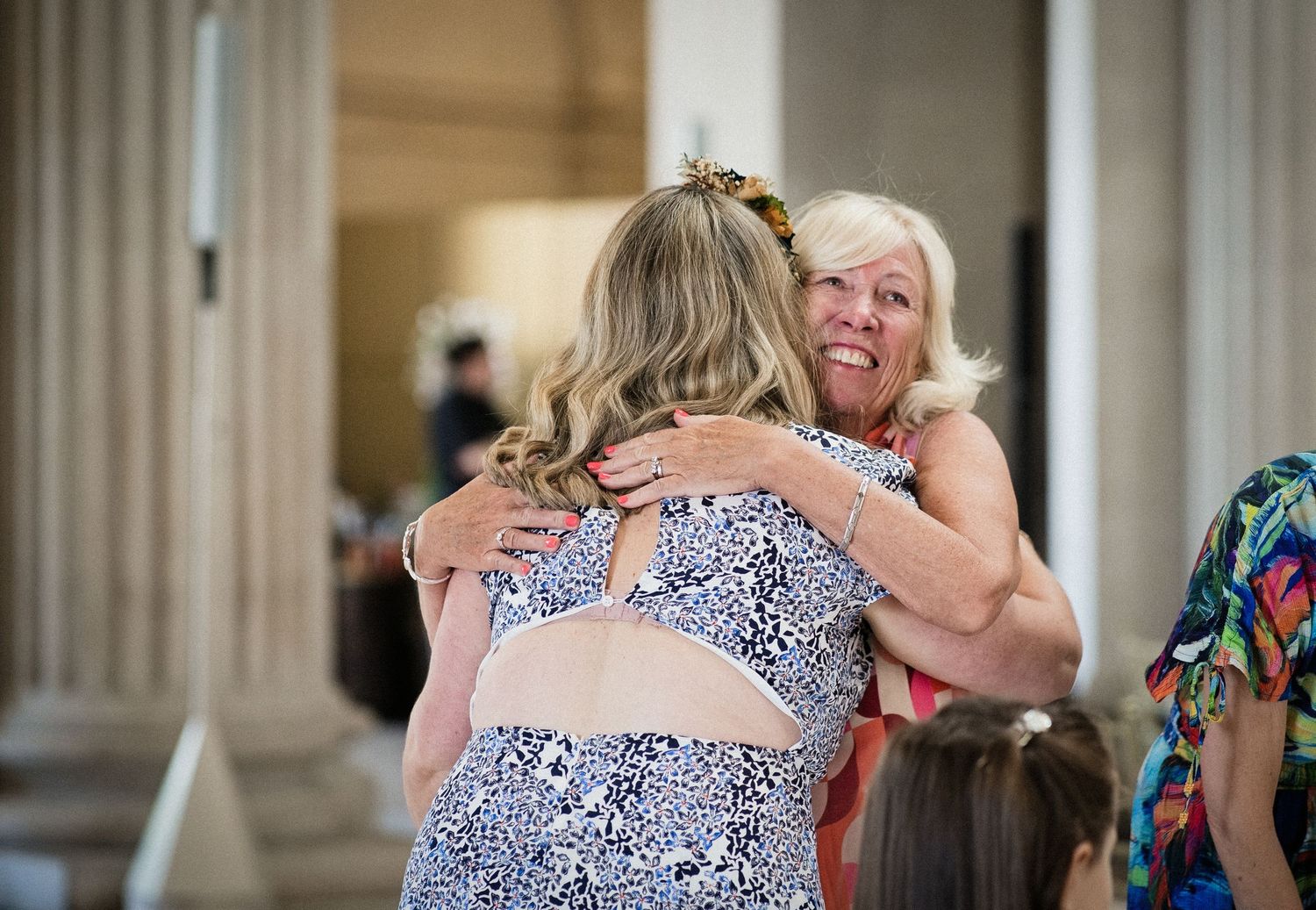 Two women share a warm embrace at an elegant columned venue during a social gathering.