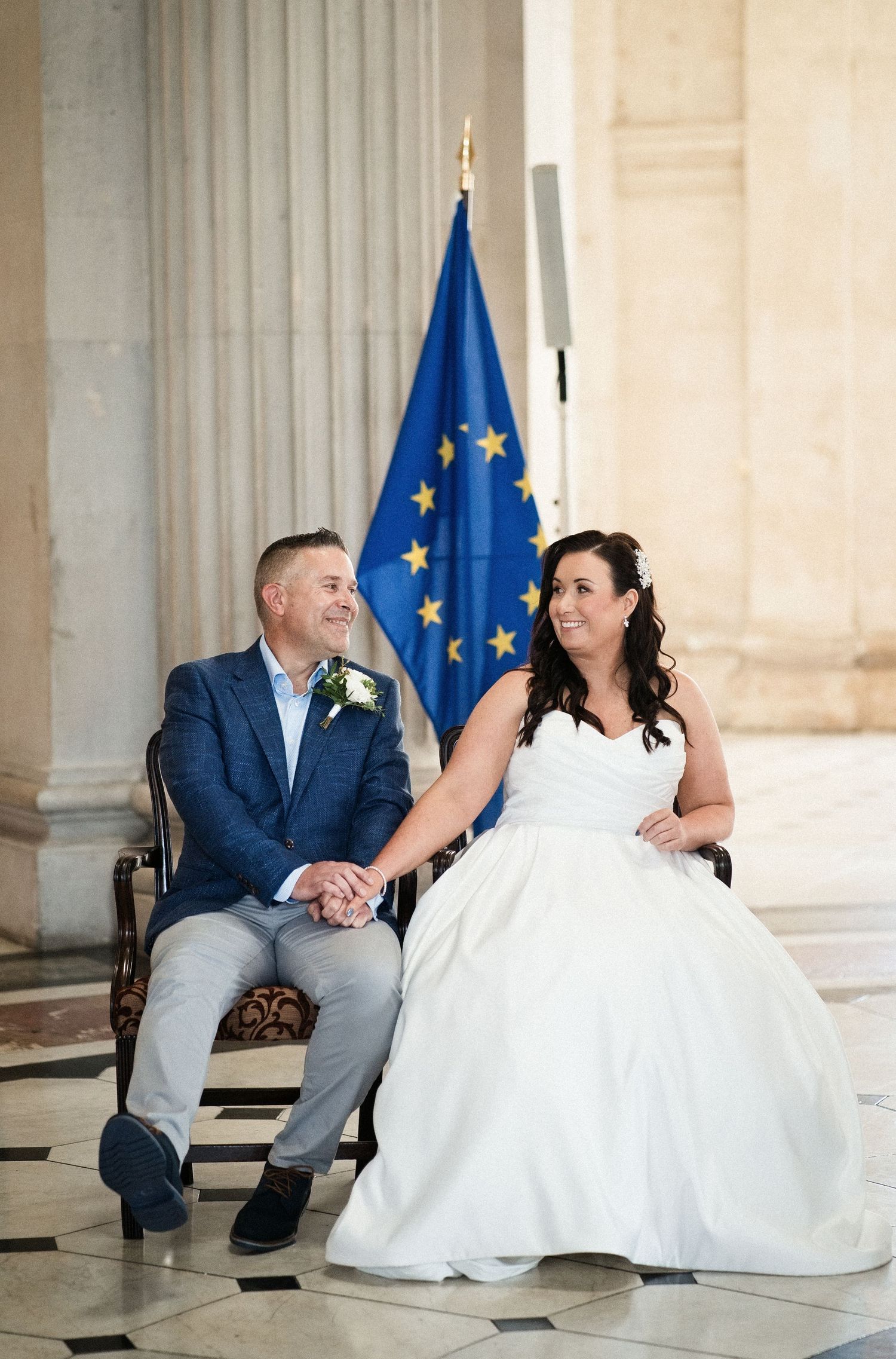 Bride in white gown and groom in blue suit hold hands seated before EU flag at wedding venue.