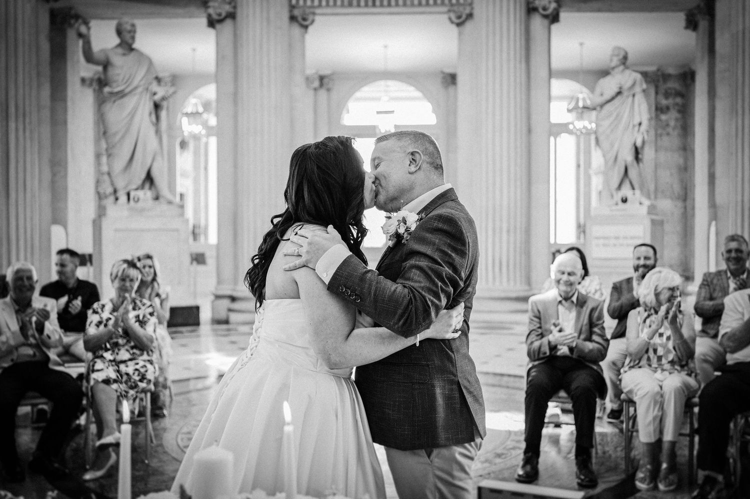 Black and white photo of a bride and groom sharing their first kiss at a wedding ceremony in an elegant hall.