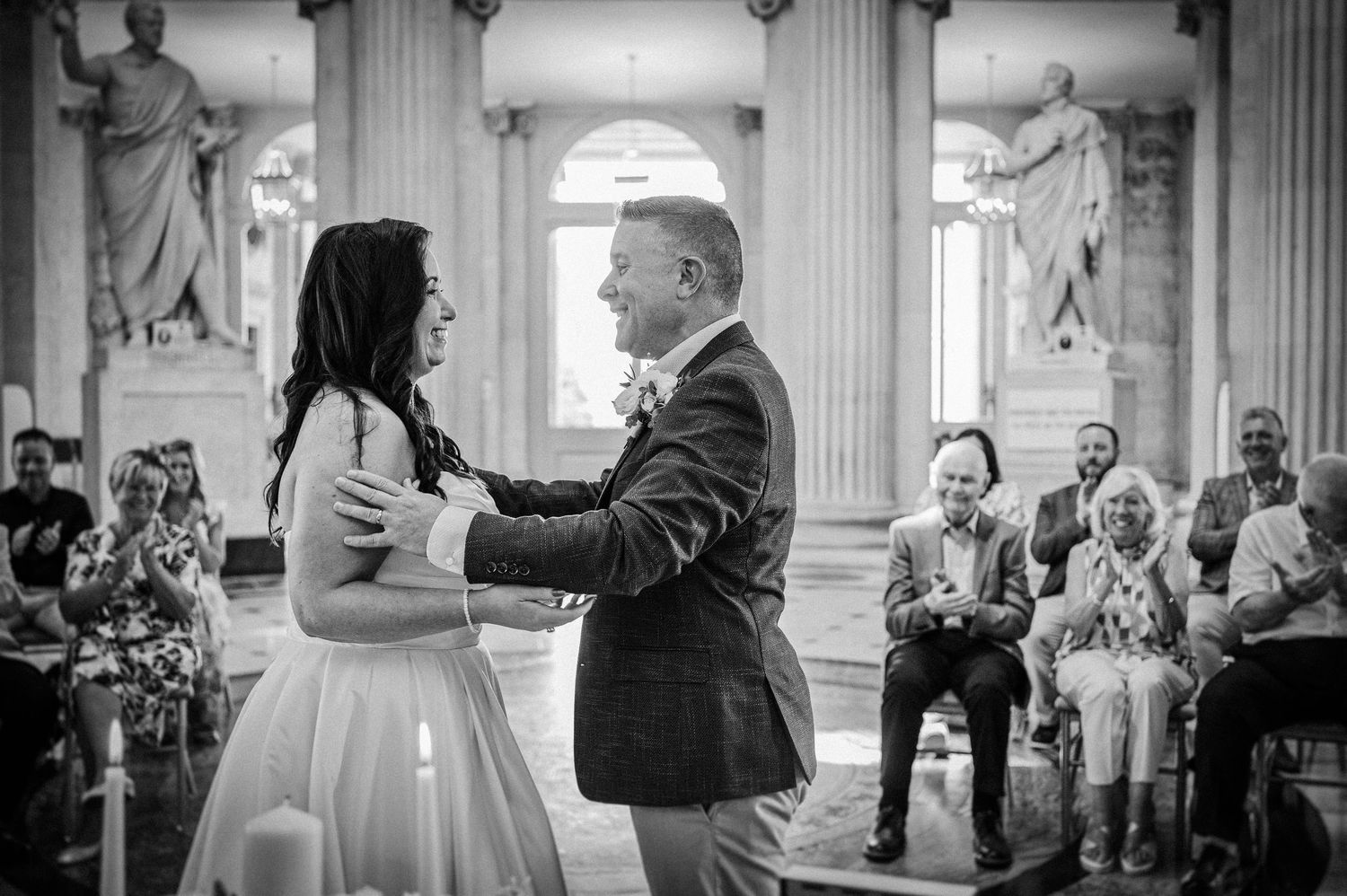 Black and white photo of bride and groom sharing first dance before guests at elegant wedding ceremony.