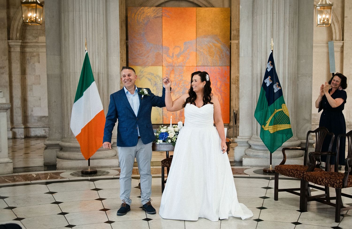 Couple celebrates wedding ceremony in elegant hall with Irish flag, bride in white gown, groom in blue suit.