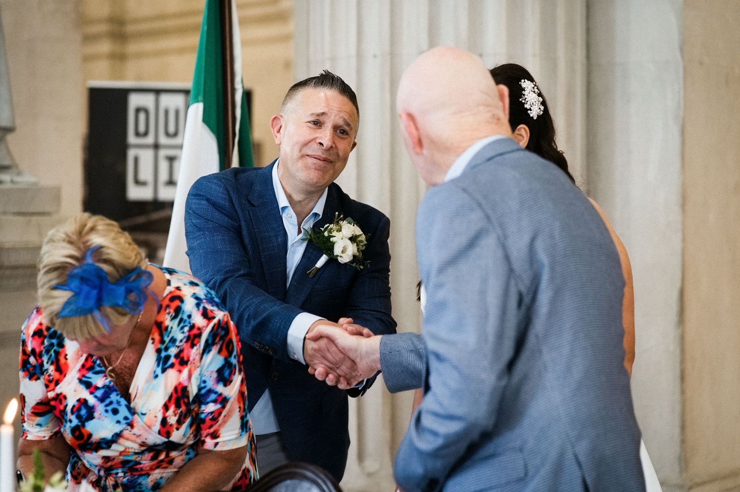 Groom in blue suit shaking hands at Dublin wedding ceremony with guests in background.