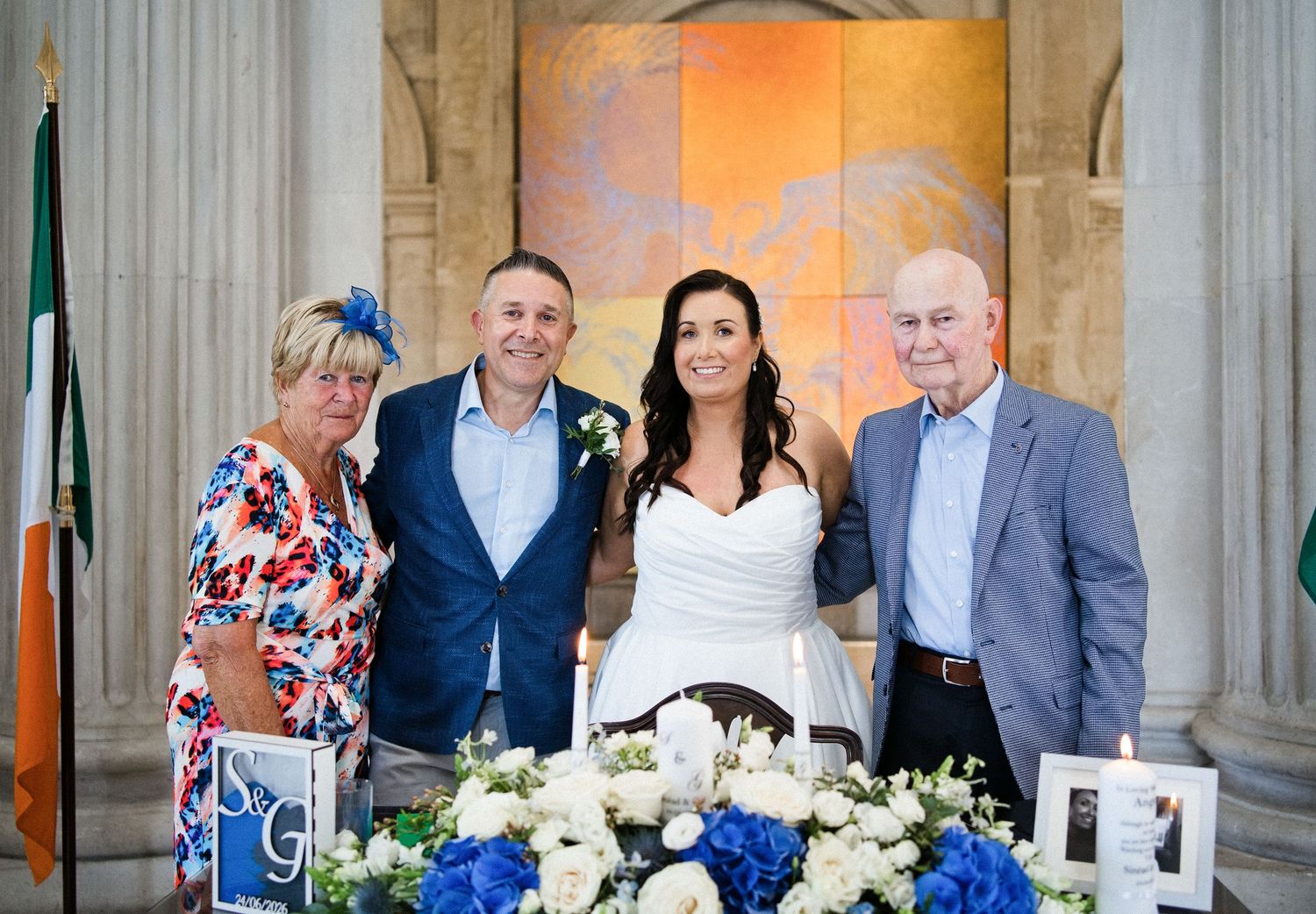 Wedding group photo with bride in white dress and three guests posing near floral arrangements with Irish flag backdrop.