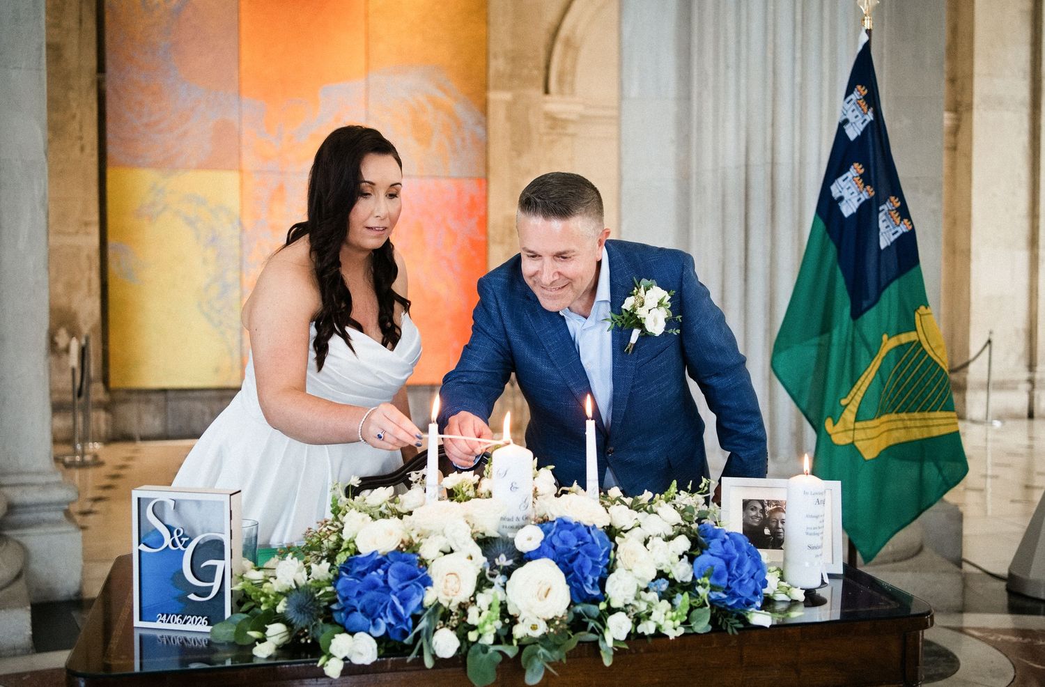 Bride and groom lighting unity candle at wedding ceremony table with blue and white floral arrangement and Irish flag.
