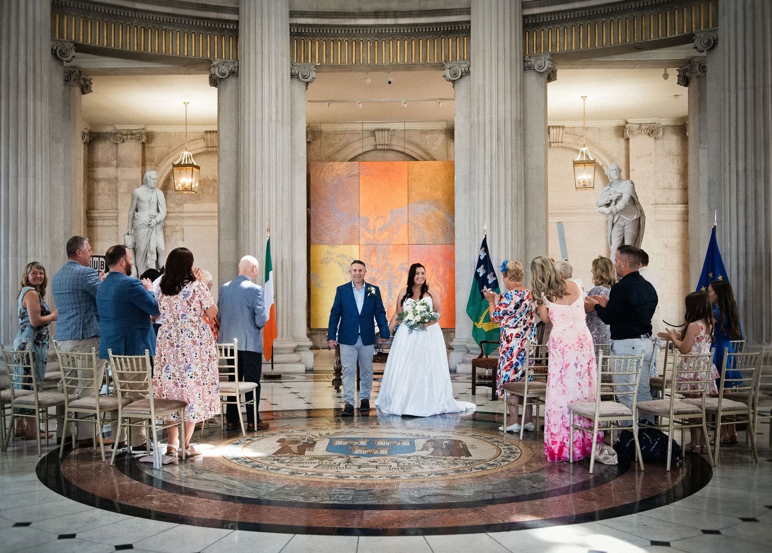 Elegant wedding ceremony in a grand columned hall with bride and groom standing before guests seated on gold chairs.